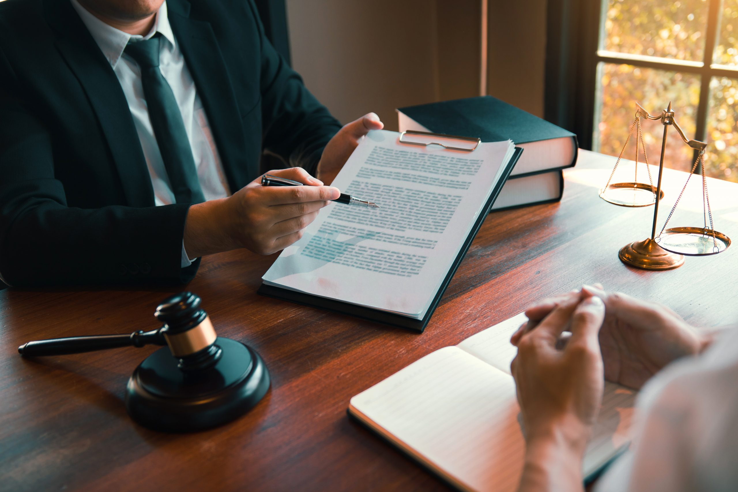 A lawyer in a suit provides client-centric legal representation as he reviews a document with a client at a wooden desk, with a gavel and legal scales visible, in a professional office setting.