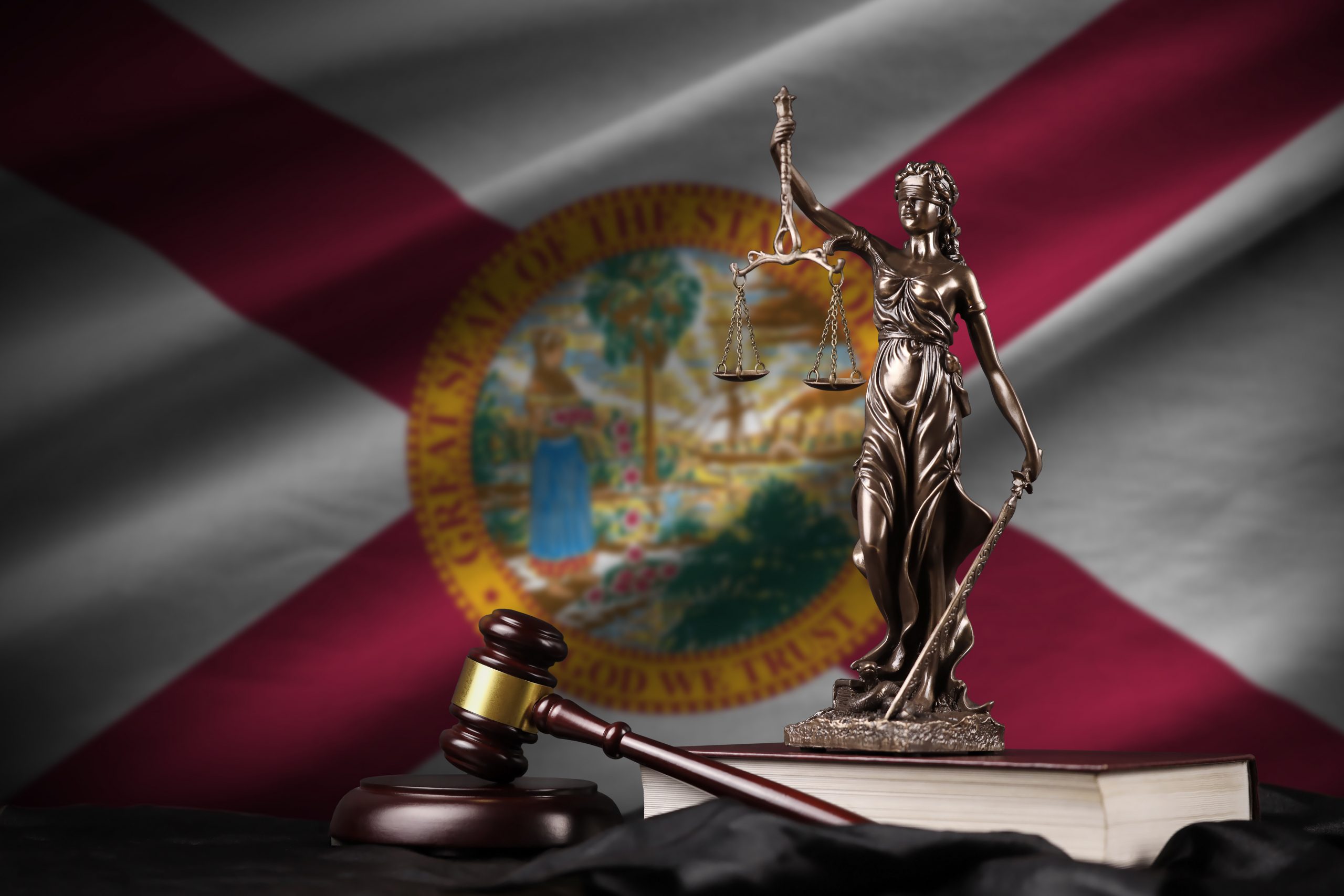 A bronze statue of Lady Justice holding scales and a sword, standing on a book beside a wooden gavel, set against a blurred backdrop of the Florida legal system flag.