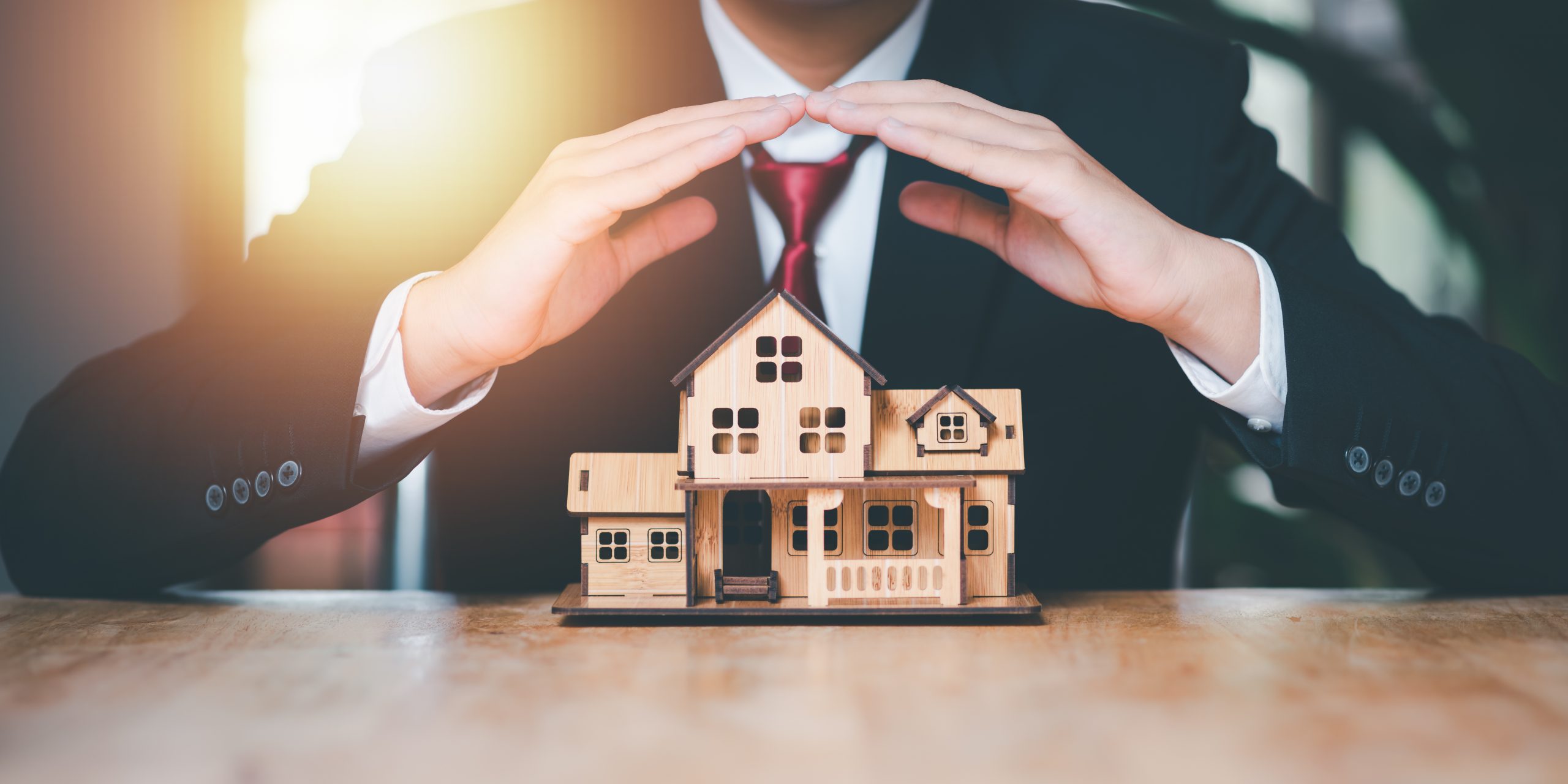 A professional in a suit making a protective gesture over a wooden house model, symbolizing insurance, security, or real estate services, specifically focusing on First Party Property Claims.