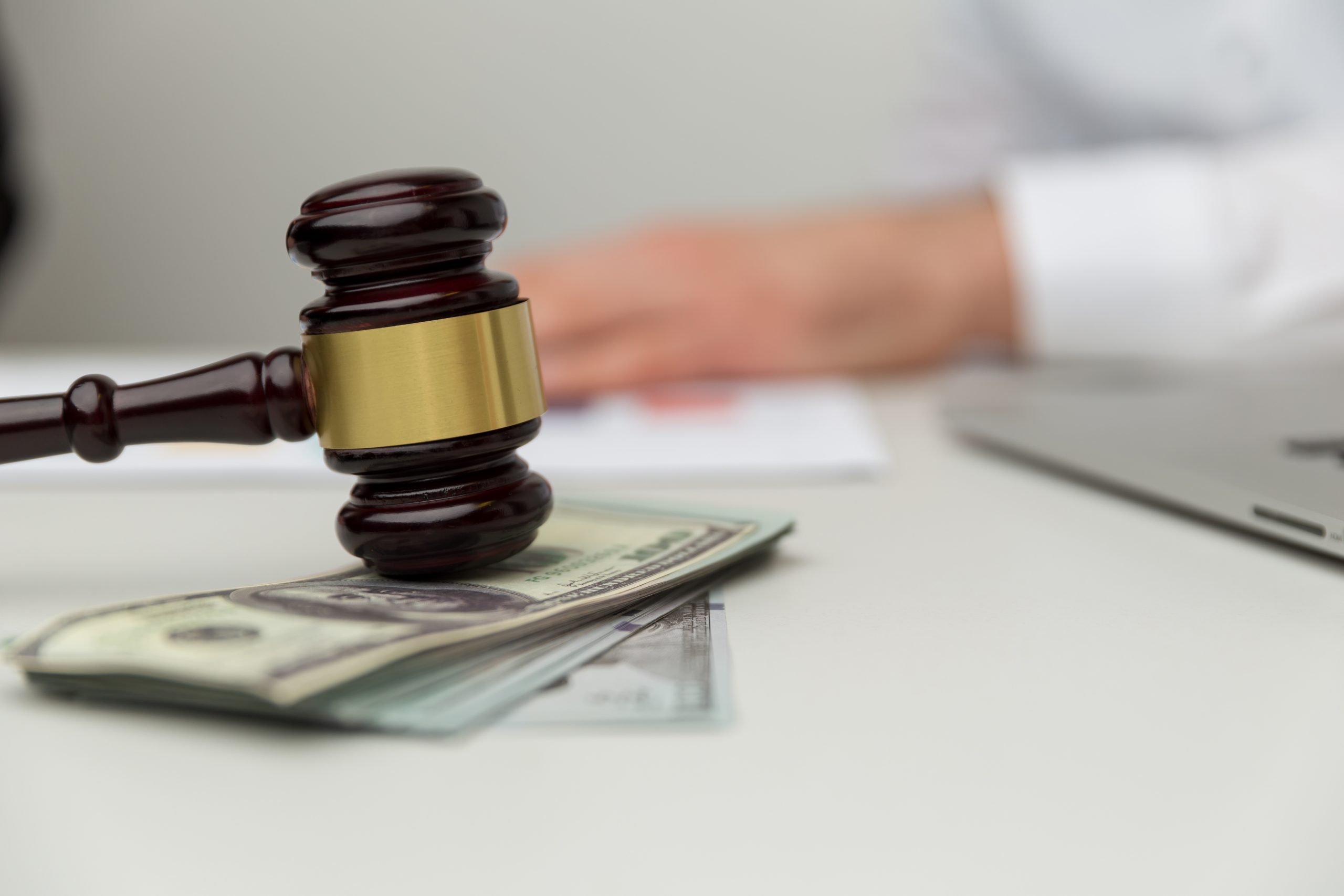 A wooden gavel with a brass band rests on top of a stack of US dollar bills on a white desk. In the background, blurred, are the hands of a person and a laptop. The scene suggests the costs and benefits of legal action in a financial context.