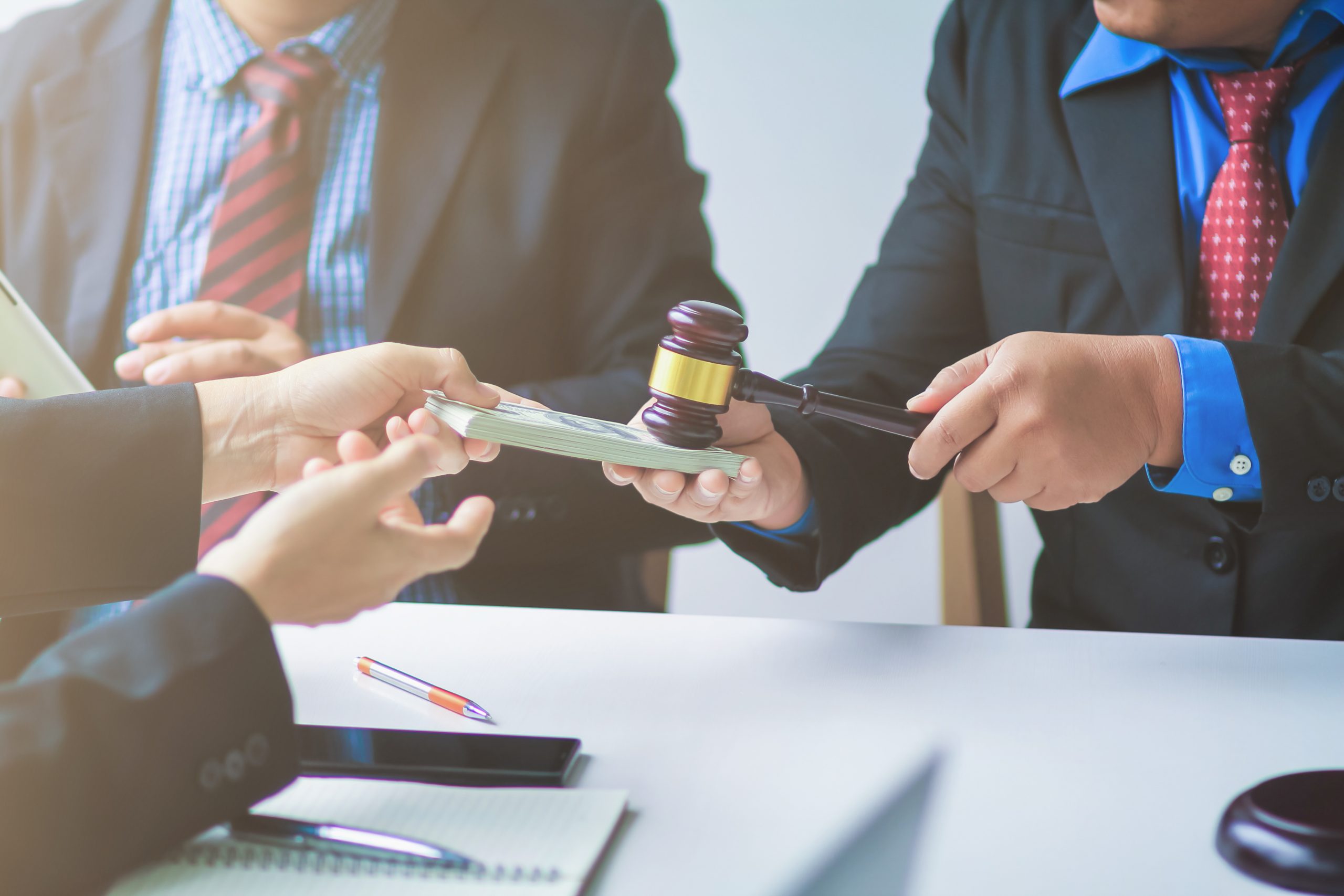 Three people in suits are seated at a table. One person, using a gavel, strikes a stack of cash held by another, symbolizing their fight against unyielding insurance companies. The third person reaches out towards the cash. Items such as a pen, a notebook, and a smartphone are visible on the table.