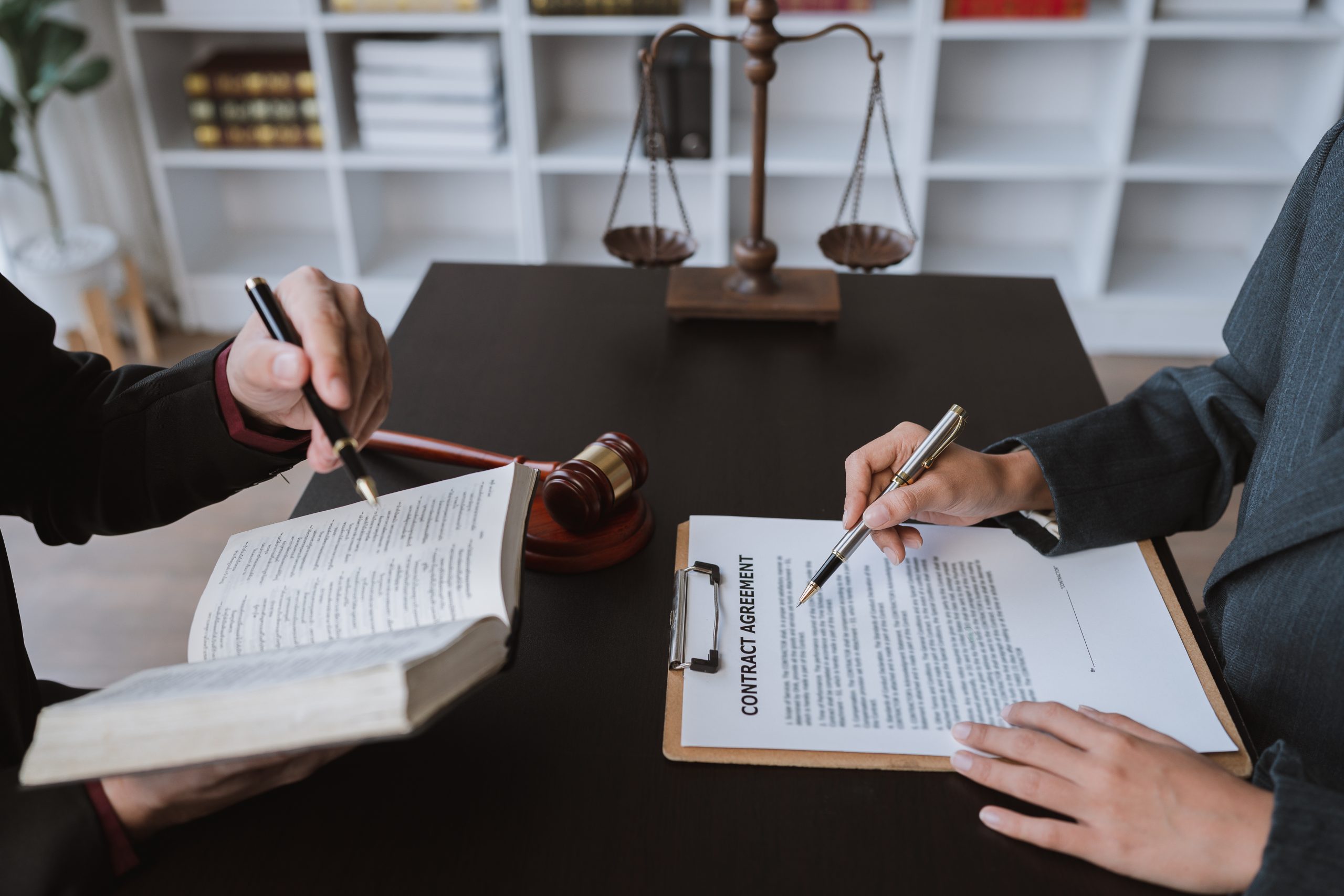 Two people sit at a desk with legal documents. One points at a contract on a clipboard with a pen, while the other holds and writes in a book. A judge’s gavel and scales of justice are visible in the background, emphasizing the importance of timely legal action to meet statutes of limitations.