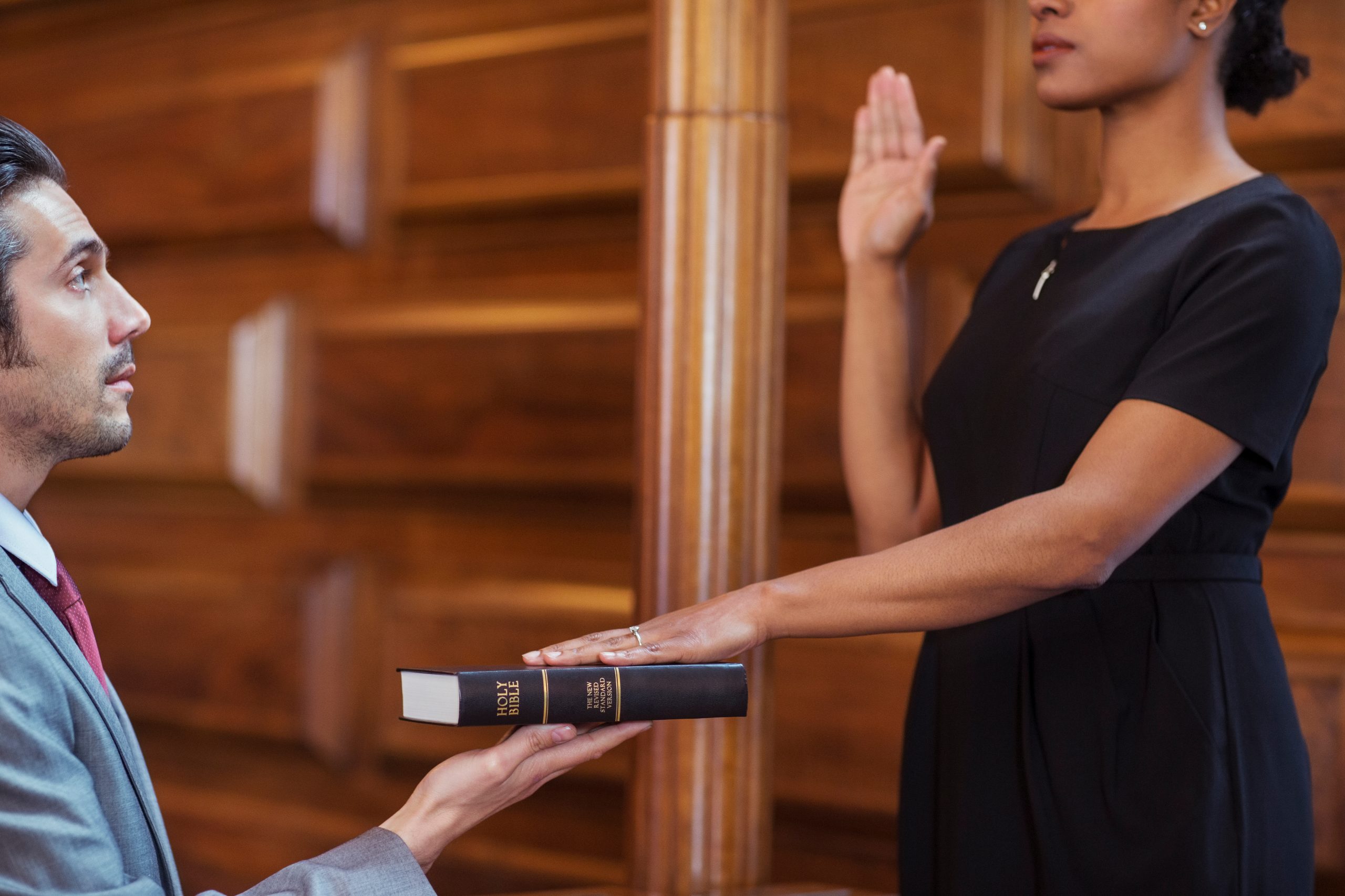 A woman raises her right hand while placing her left hand on a Bible held by a man. The scene takes place in a wood-paneled room, likely a courtroom, indicating an oath or swearing-in ceremony, possibly involving expert witnesses in significant legal cases.