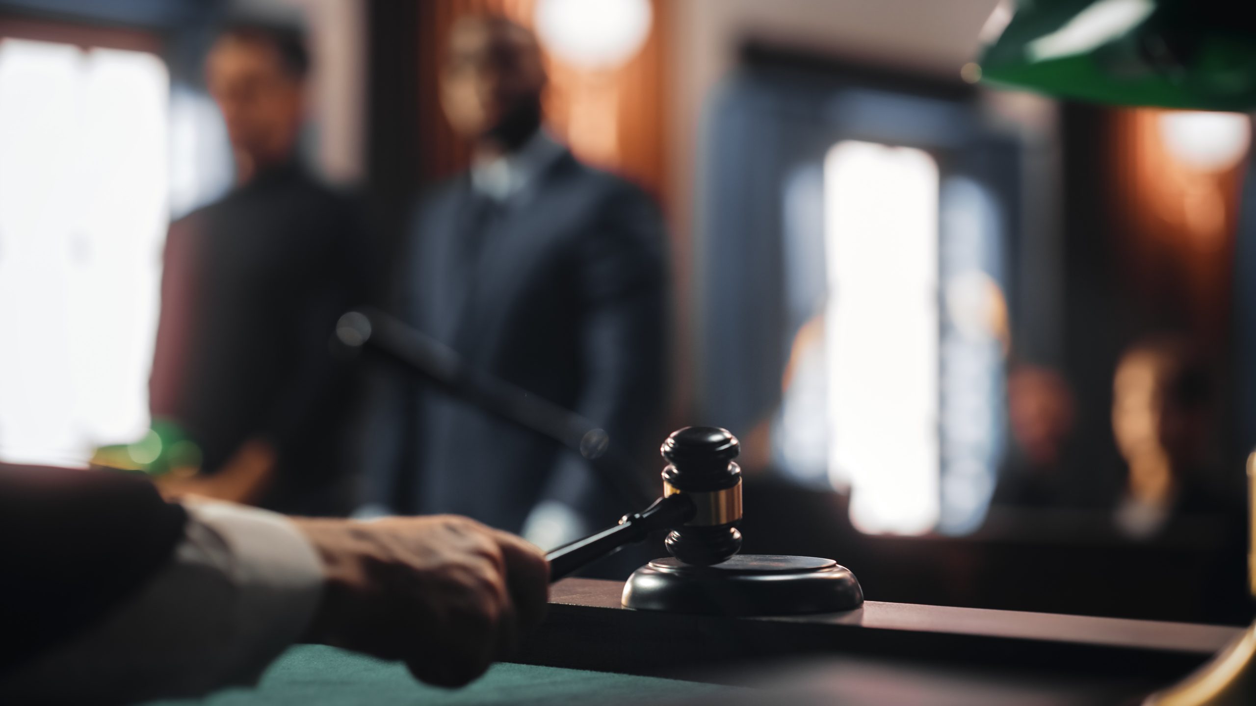 Close-up of a judge’s gavel being struck in a courtroom. In the blurred background, two individuals stand facing the judge, possibly going to trial. The scene is dimly lit, creating a serious and formal atmosphere.