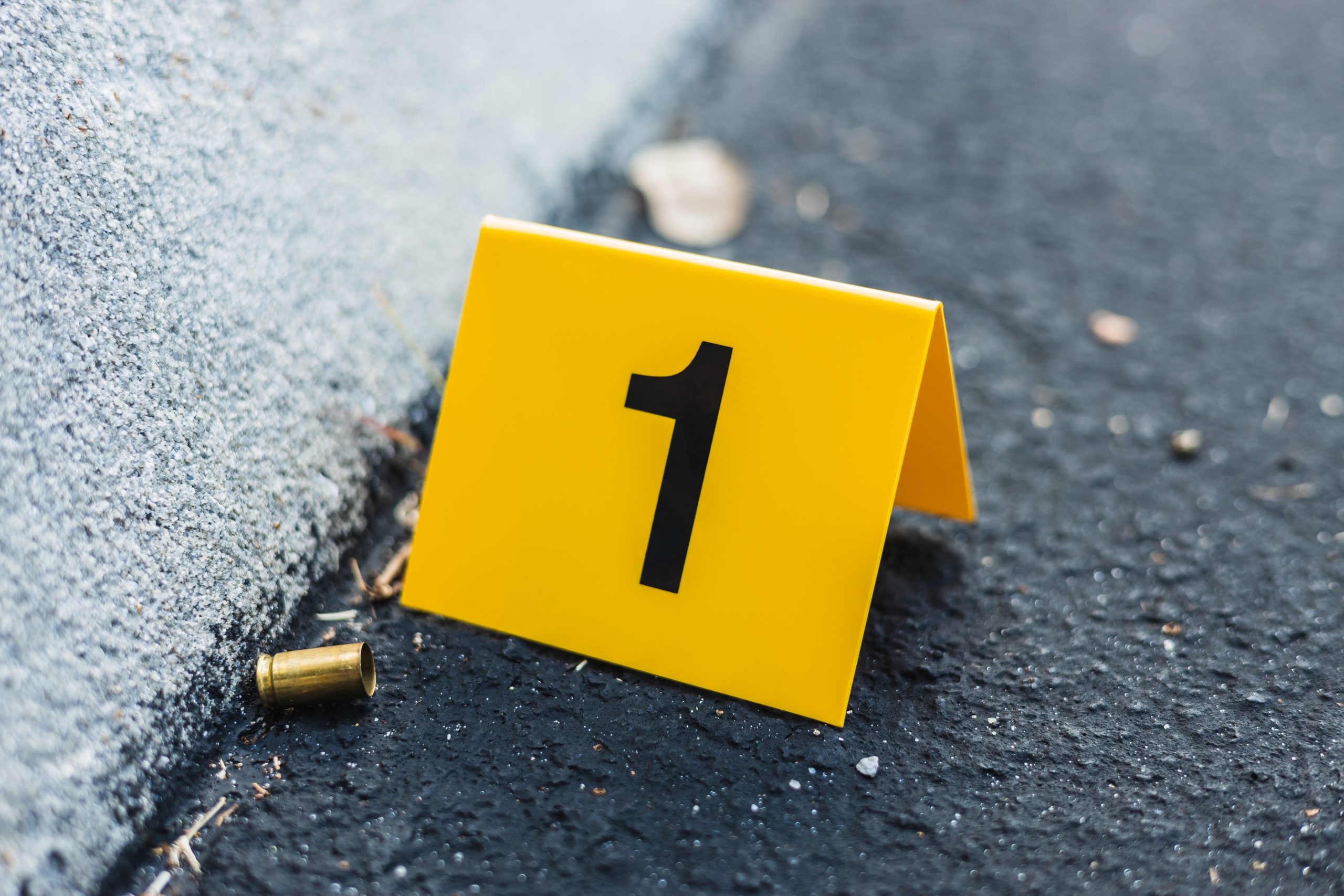 A yellow evidence marker with the number “1” stands next to a bullet casing on an asphalt road, indicating a crime scene detail. The marker is placed near the curb of the road, potentially relevant in personal injury cases involving gunfire incidents.