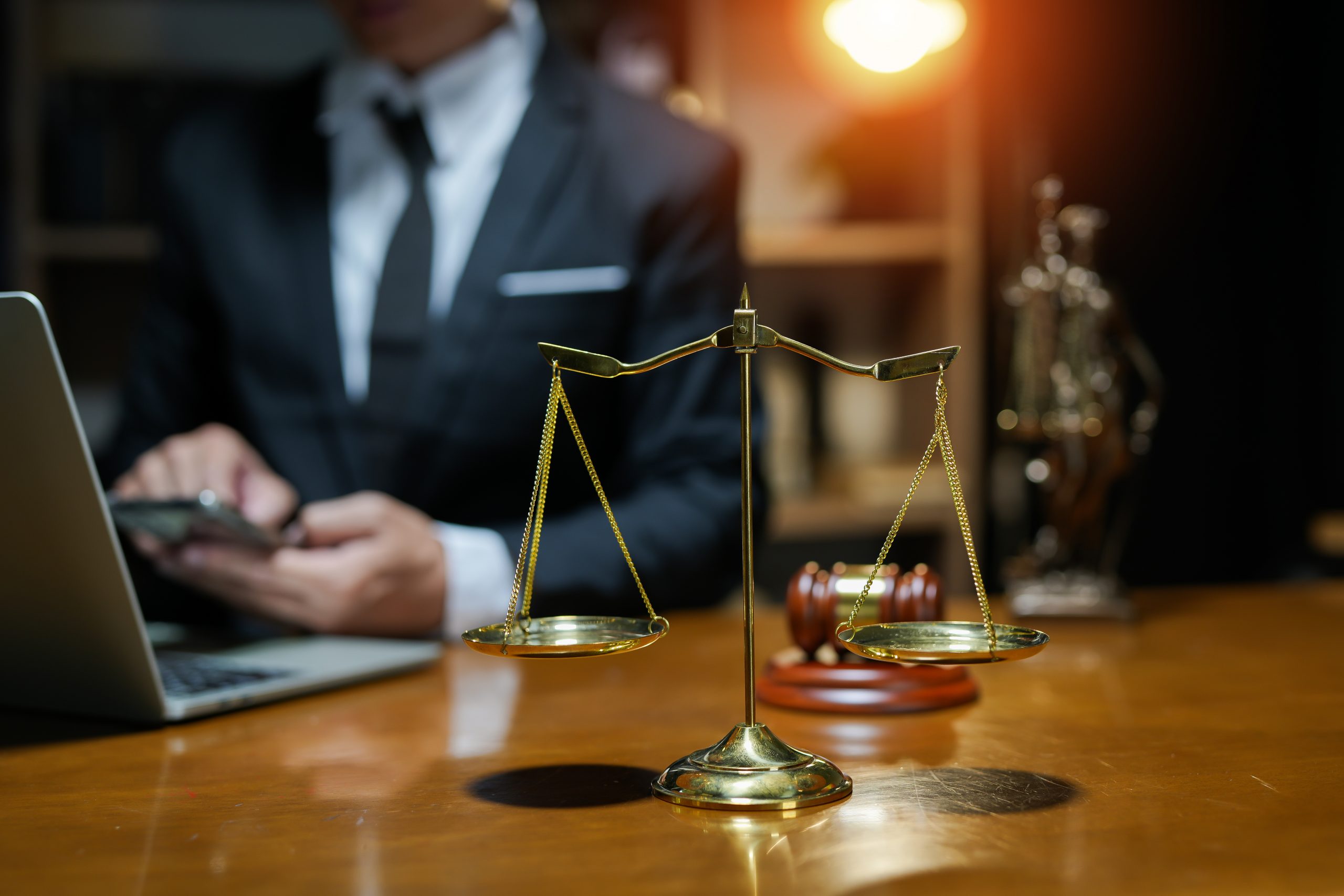 Close-up of a golden balance scale on a wooden desk, with a blurred person in a suit typing on a laptop in the background. A gavel and legal statue are also visible on the desk, suggesting an office where experienced attorneys work diligently to provide justice.