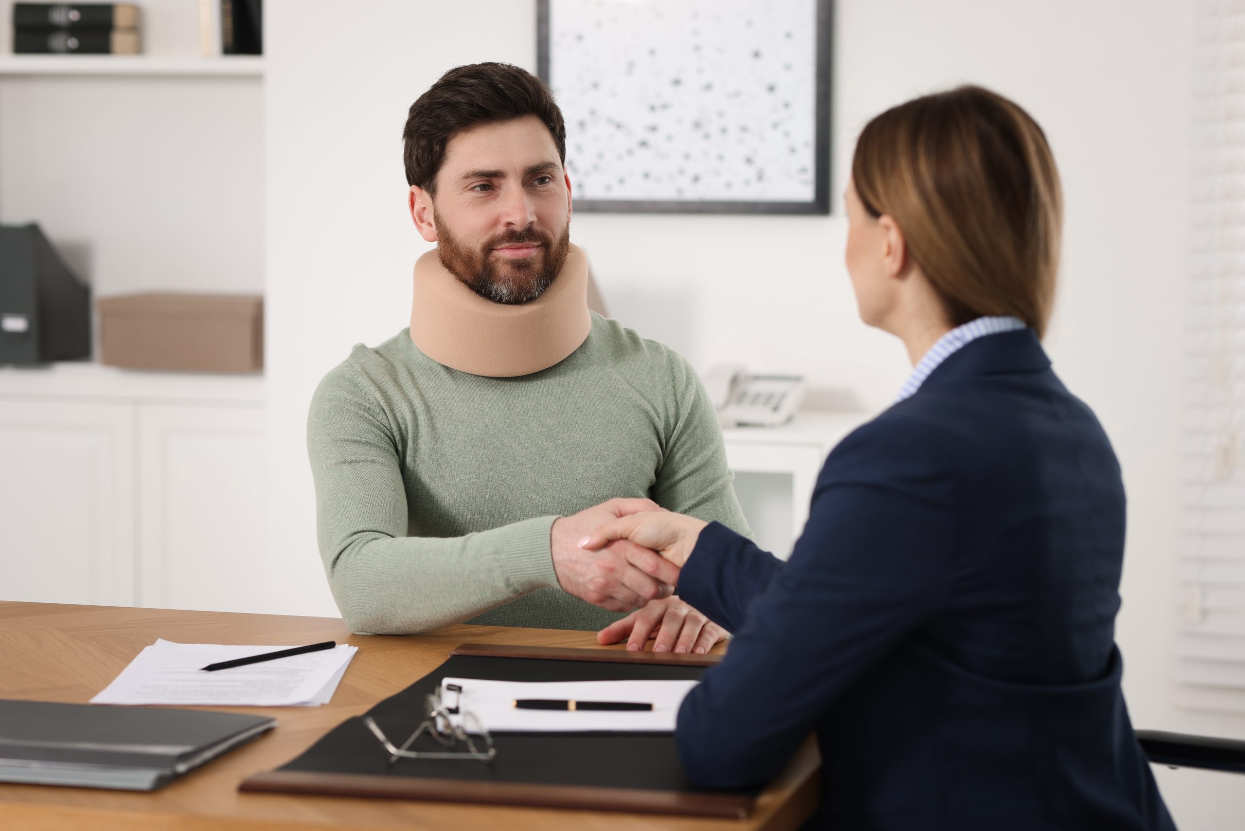 A man wearing a neck brace is sitting at a desk, shaking hands with a woman in a blue jacket. Papers and a clipboard are on the desk in front of them, suggesting a consultation for legal advice regarding his traumatic injuries. They are in a bright office setting.