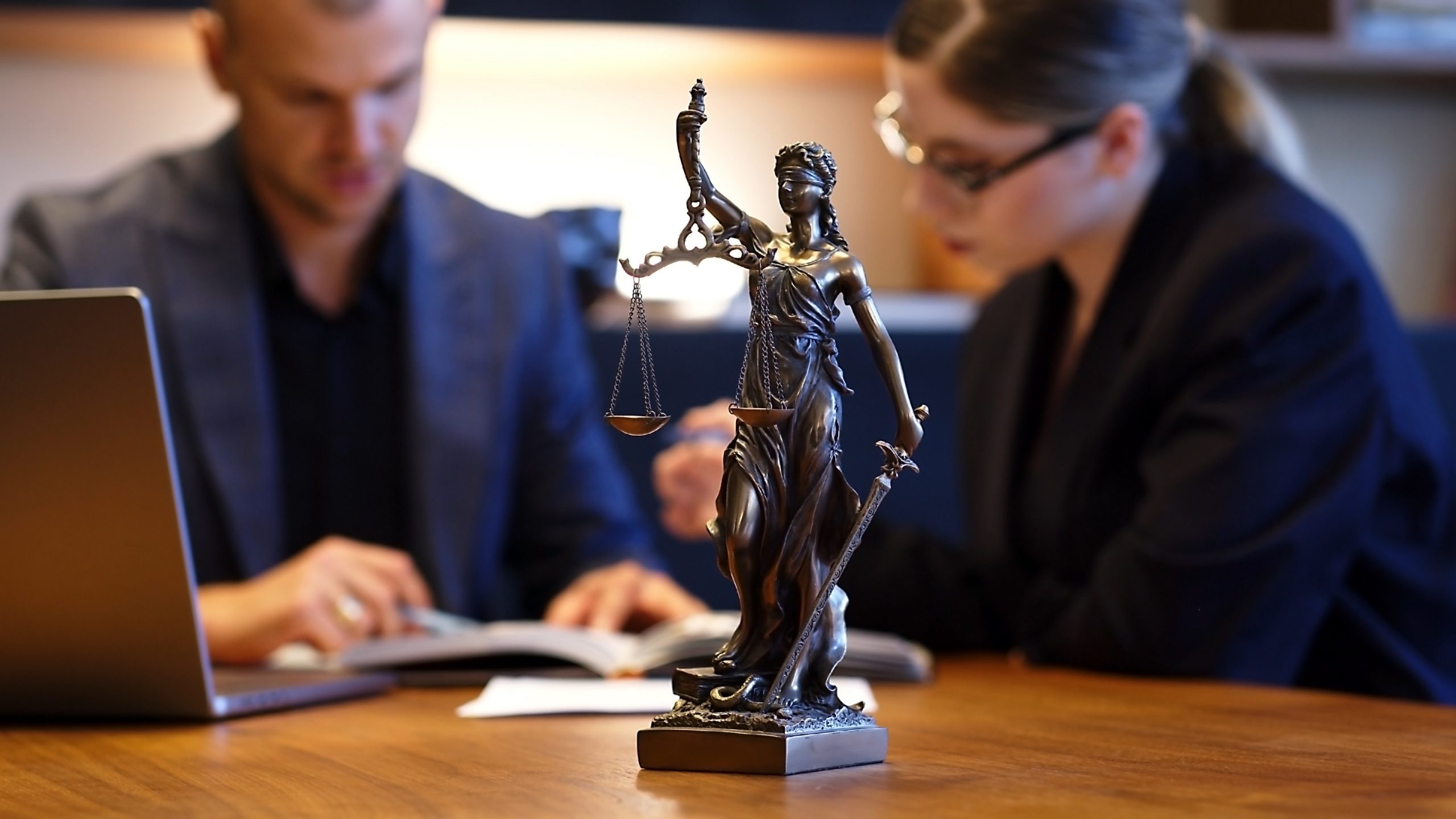 A bronze statue of Lady Justice is prominently displayed on a wooden table. In the background, a man and a woman, both dressed in professional attire, are engaged in a discussion over open books. The room, designed as a primer for the legal system to laypersons, is warmly lit.