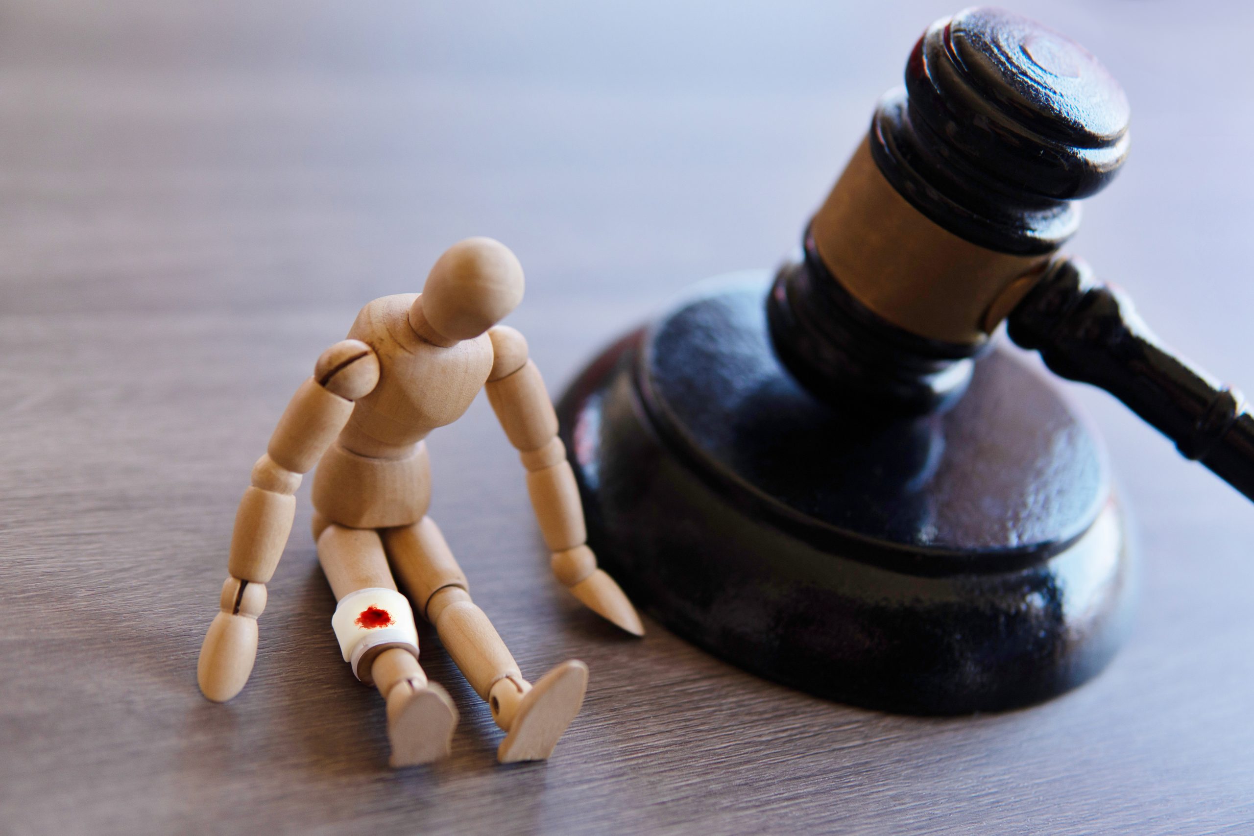 A wooden artist mannequin with a bandage on its knee, displaying a red mark, sits leaning against a large gavel on a wooden surface, symbolizing injury and legal justice, representing the varied types of personal injury cases.