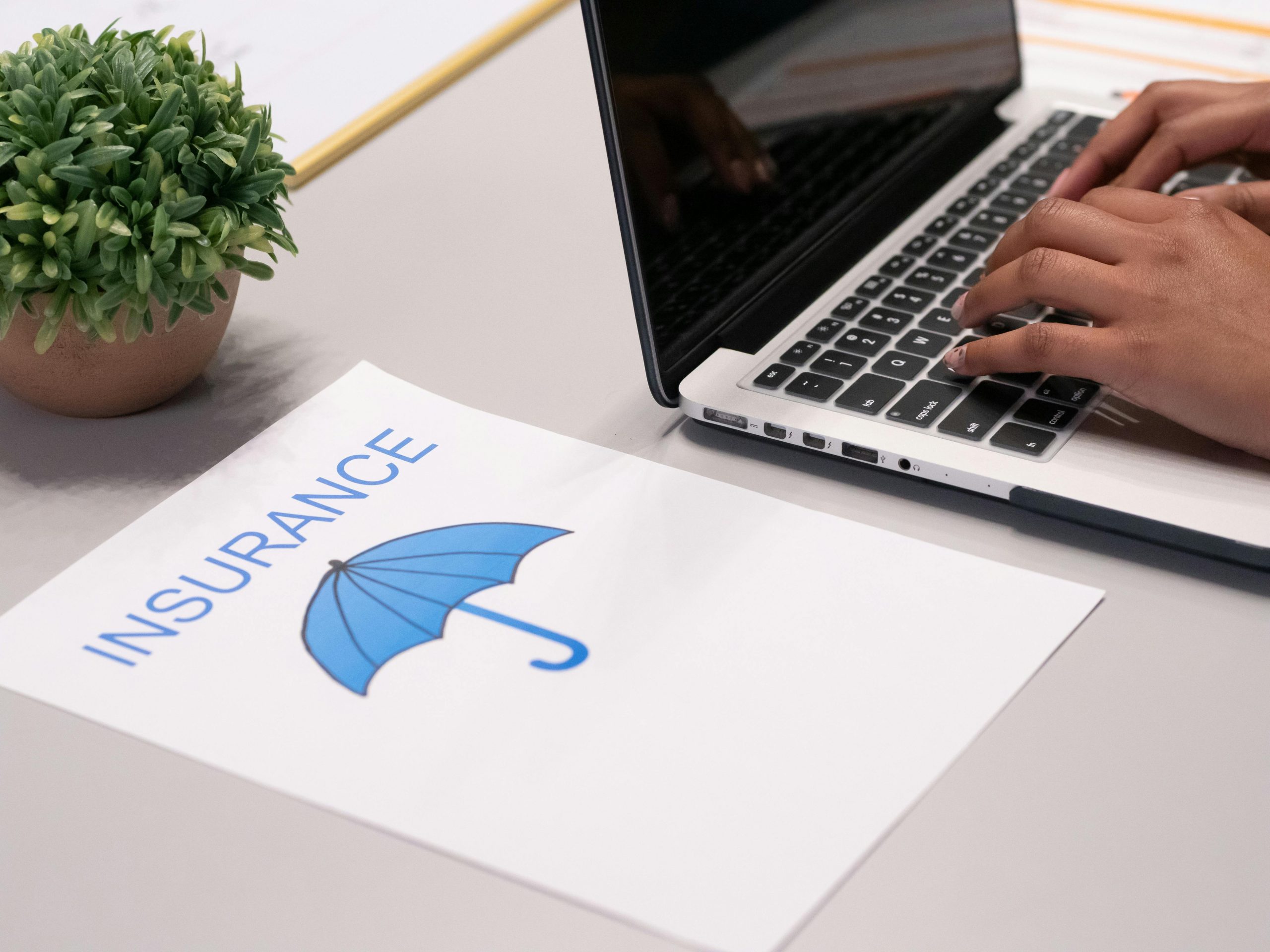 A person’s hands are typing on a laptop keyboard. Next to the laptop is a white paper with the word “INSURANCE” and a blue umbrella logo, highlighting personal injury protection. There is also a small potted plant on the desk.