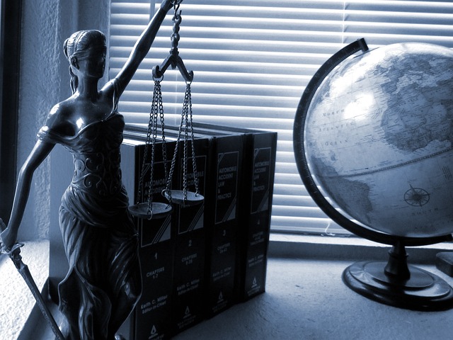 A statuette of Lady Justice stands beside a stack of thick books and a globe on a windowsill, embodying the spirit of litigation. The statuette holds scales and a sword, symbolizing law and justice. Blinds cover the window in the background, casting soft light on the objects.