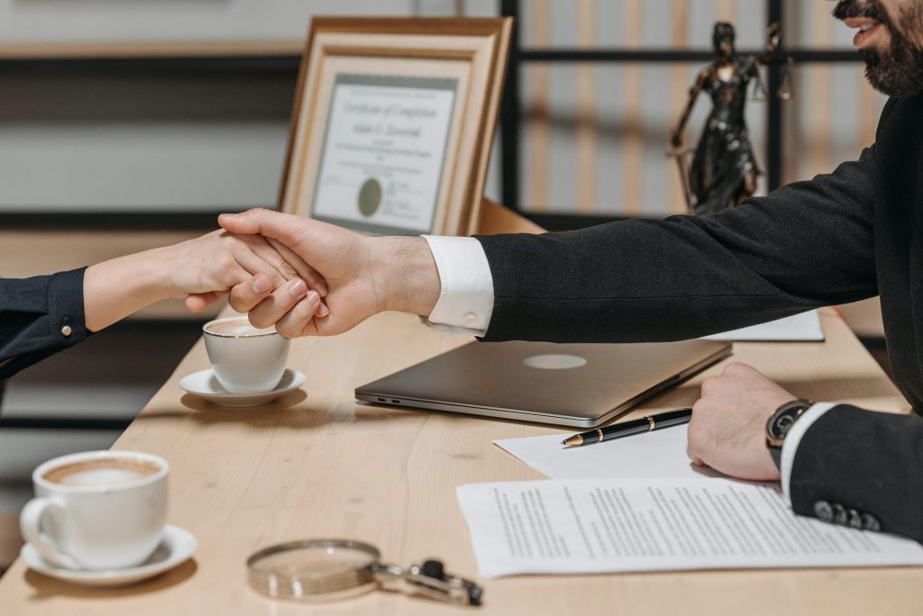 Two people are shaking hands across a desk in an office setting, suggesting the conclusion of a successful litigation. The desk has coffee cups, a laptop, a pen, papers, a magnifying glass, and a framed certificate. A statuette of Lady Justice is visible in the background.