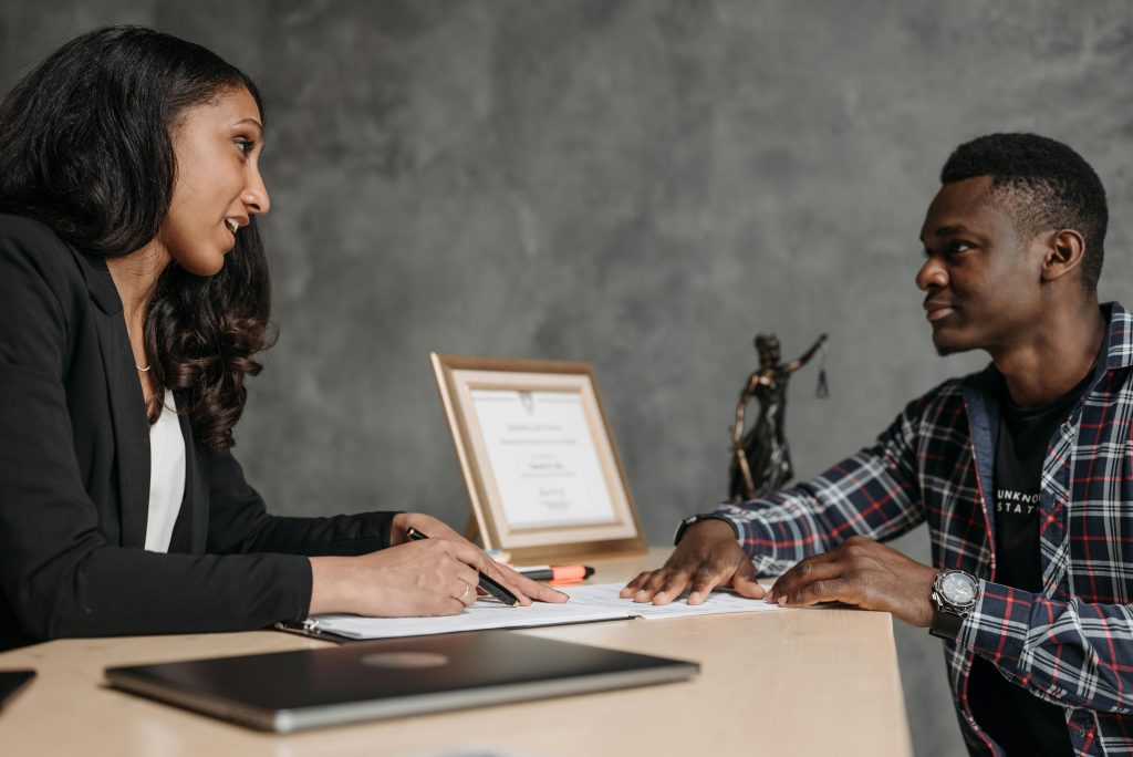 A woman and a man sit across from each other at a desk. The woman, holding a pen, looks at the man, who is listening attentively. On the desk lies a laptop, documents, and a framed certificate beside a Lady Justice statue—a scene embodying the principles of client advocacy.