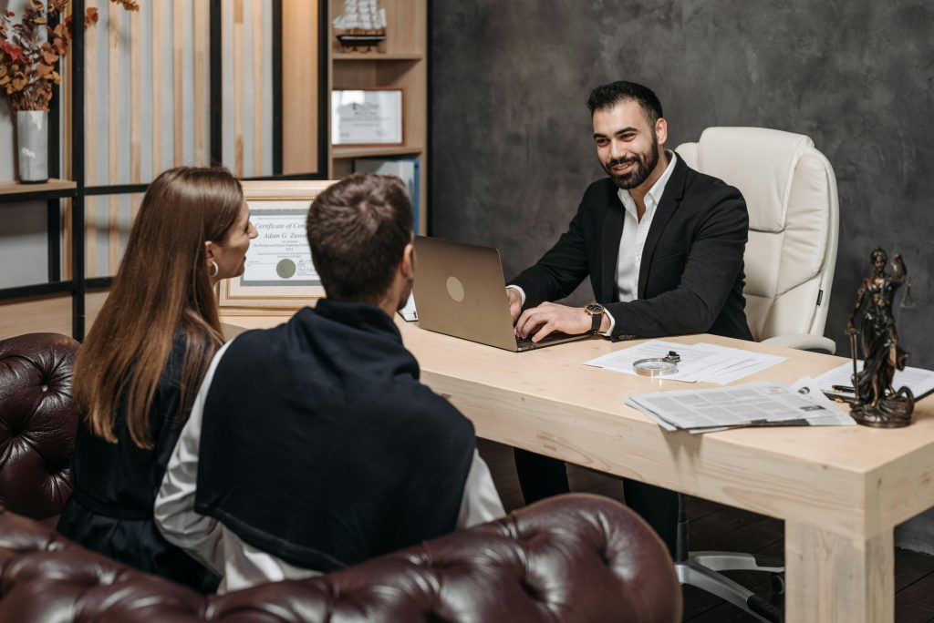 A man in a suit sits at a desk with a laptop, smiling and discussing client advocacy with a couple seated across from him in a modern office. The desk has paperwork, a newspaper, and a small statue of Lady Justice. Shelves with certificates and ornaments are in the background.