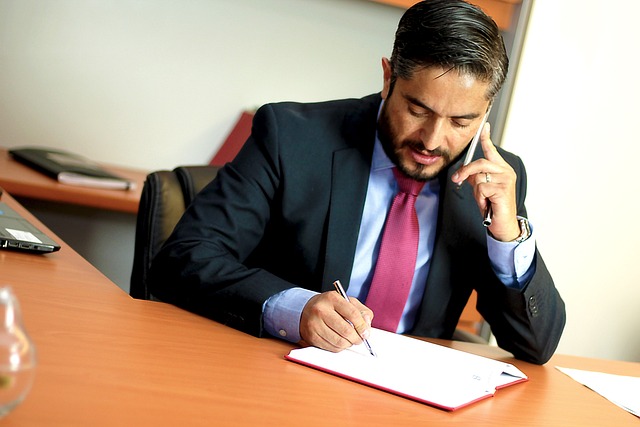 A man with dark hair and a beard, wearing a blue suit and red tie, is seated at a wooden desk. He is talking on a cell phone with his left hand, passionately discussing client advocacy, while writing in a white notebook with his right hand. A blurred office setting is in the background.