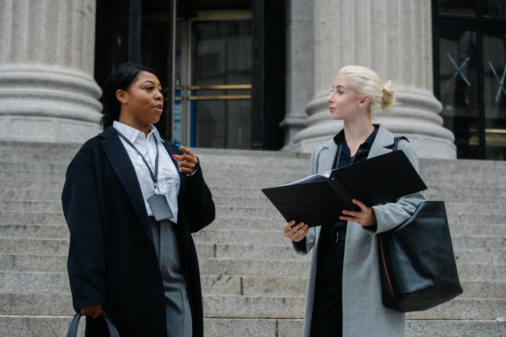 Two women in professional attire stand on steps outside a stone building with large columns. The woman on the left is speaking and gesturing with her hand, while the woman on the right holds a litigation folder and listens attentively. Both carry bags.