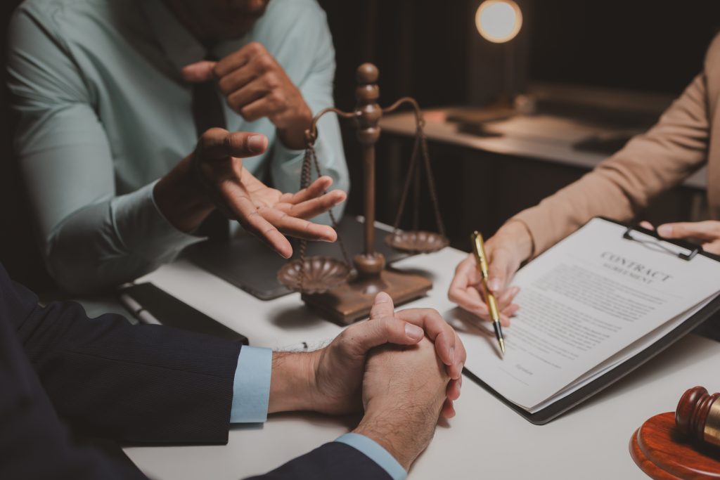 Three people are seated at a table discussing a contract. One person, possibly the best attorney among them, is holding a clipboard with a contract on it and pointing to it with a pen. There is a set of scales and a gavel on the table, suggesting a legal or business meeting about personal injuries.