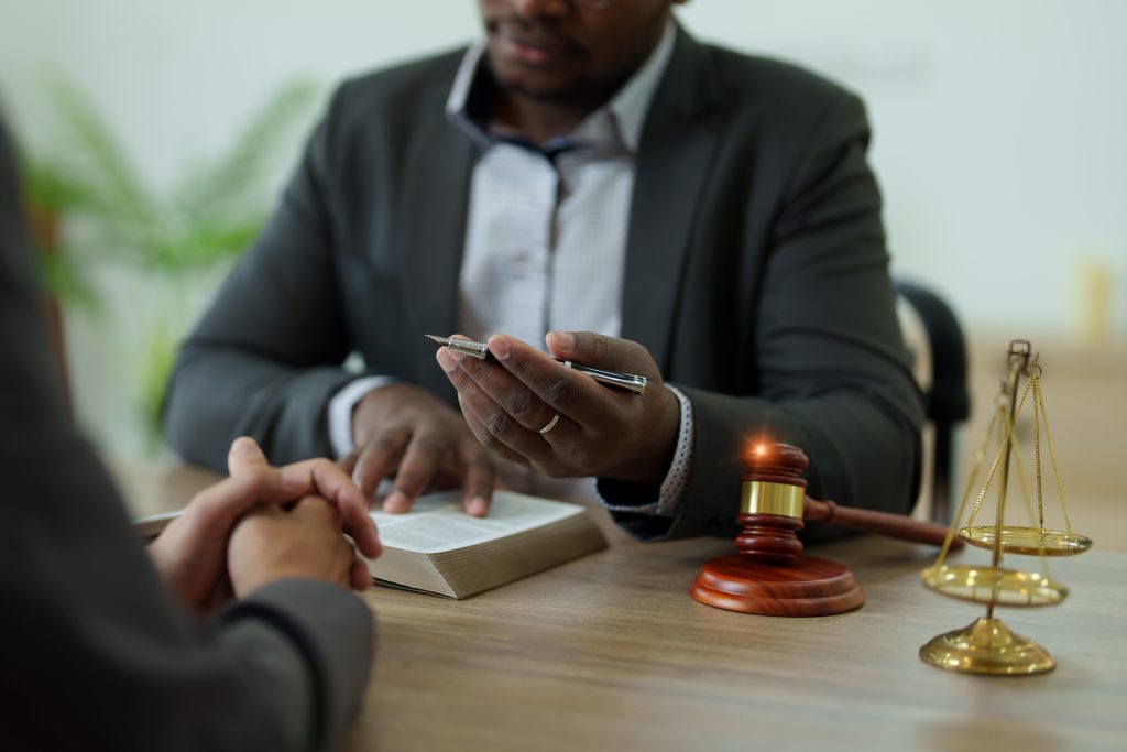 A person in a suit holds a pen and points at a book while sitting across from another individual. On the table between them are a wooden gavel and a set of scales, suggesting they might be discussing legal assistance for expungements, possibly in an attorney's office.