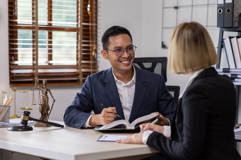 A man in a dark suit and glasses is smiling while talking to a woman with blonde hair in a business suit. They are both seated at a desk with open books, legal scales, a gavel, and stacks of files in the background—discussing top personal injury matters.