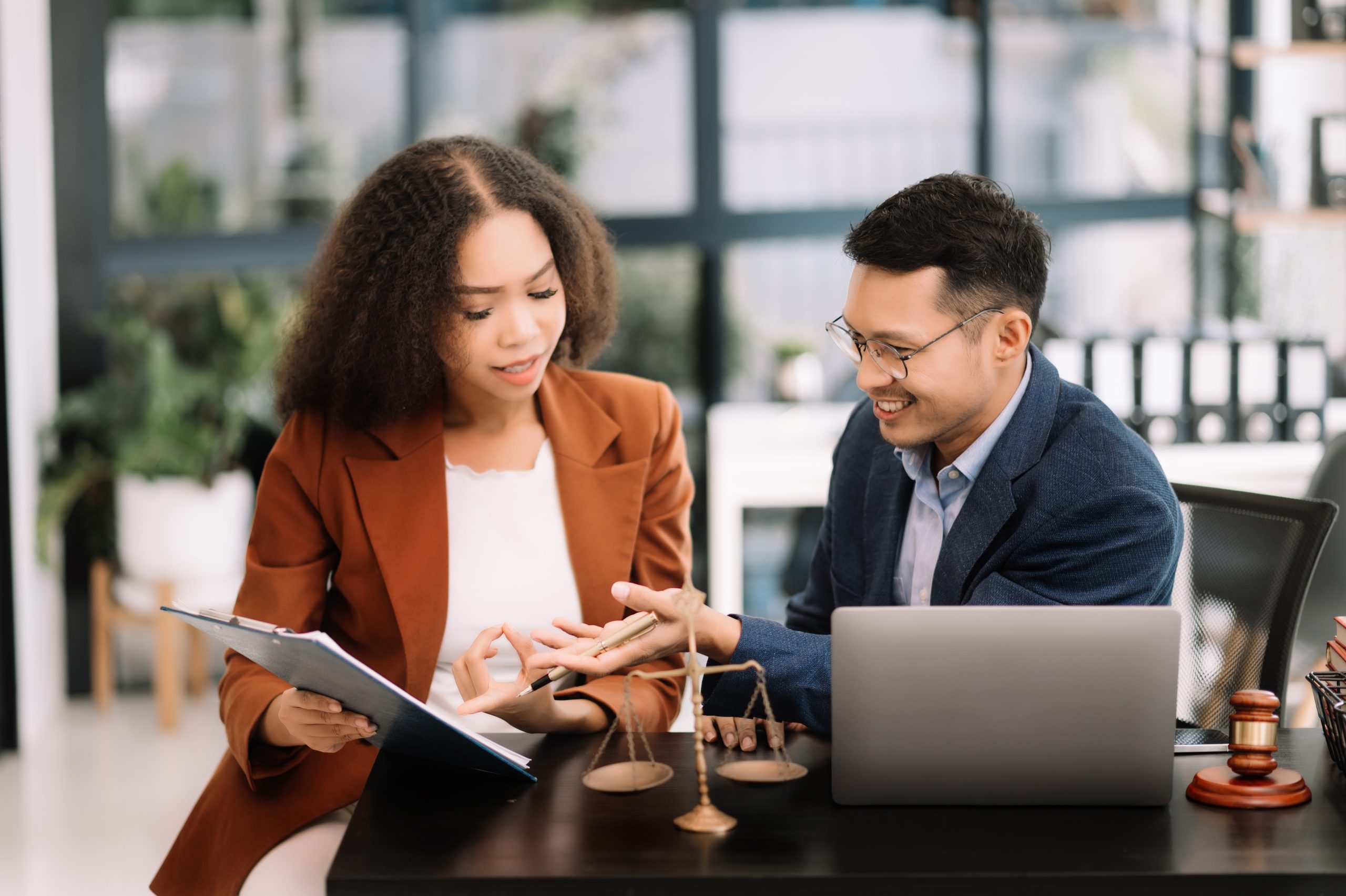 Two professionals, a woman and a man, are seated at a desk in an office. The woman, acting as the guide, is holding a clipboard and pointing to a document about expungement in Florida. The man is smiling and holding a pen. There is a laptop, scales, and a gavel on the desk.