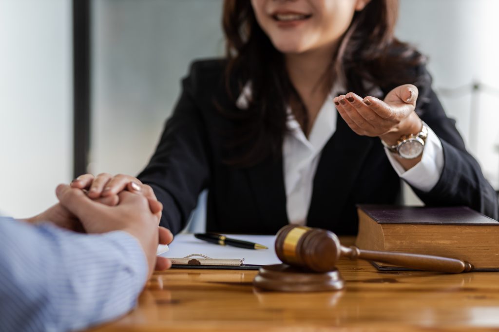 A person in a suit is sitting at a desk, gesturing and speaking to another individual, whose hands are visible. On the desk, there is a gavel, a book, and a pen, indicating a legal or courtroom setting—like consulting with the best attorney for personal injuries.