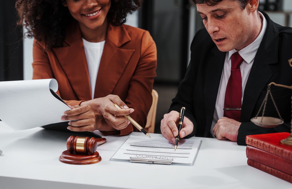 Two individuals, one with curly hair in an orange blazer and the other in a suit and red tie, sit at a desk. They are reviewing documents together, possibly discussing an expungement case. The desk has a gavel and a set of scales on it, indicating a legal setting in Florida.