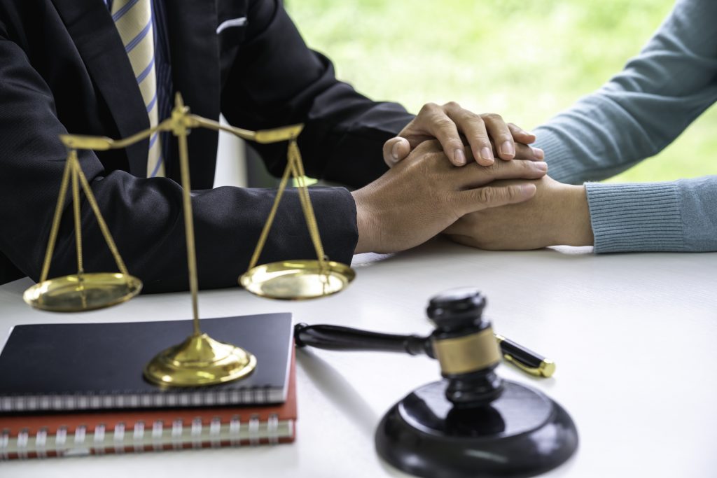 A person wearing a suit and holding a gavel in front of legal scales offers a comforting gesture by placing their hand over someone else's hands. The scene suggests attorneys providing supportive legal assistance in a professional setting.