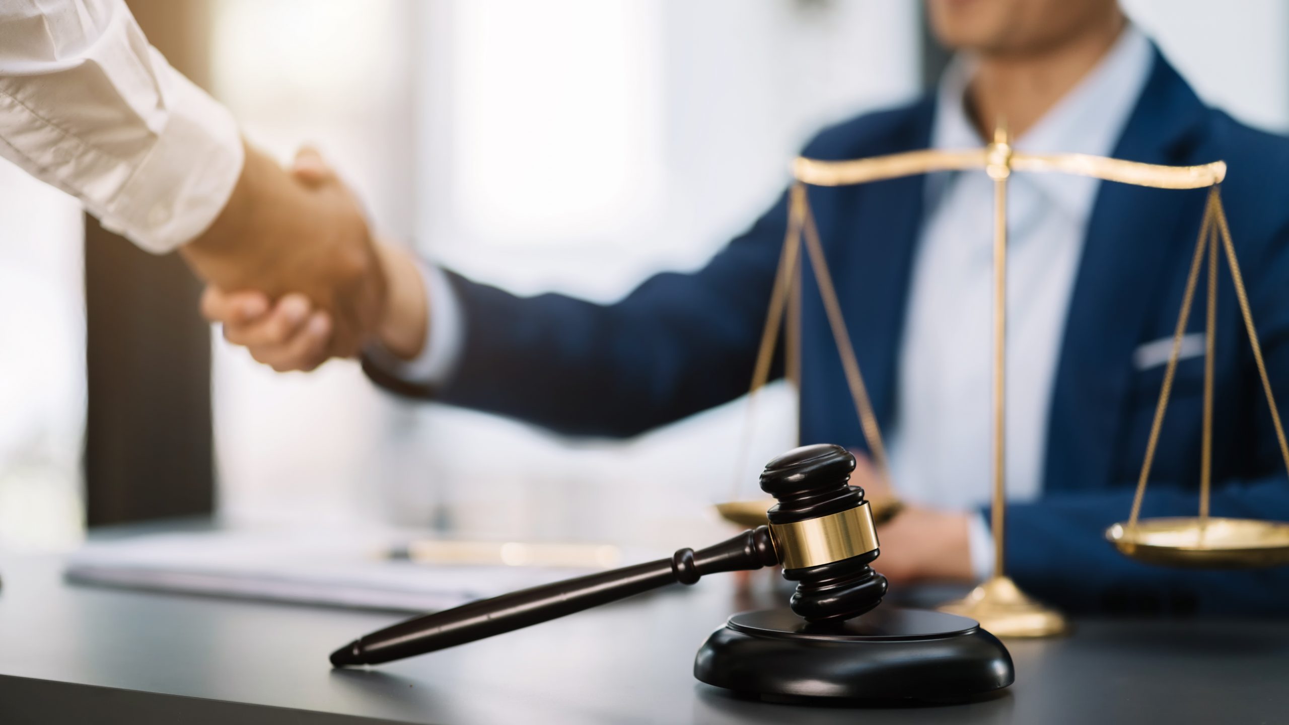 A person in a suit is shaking hands with another individual, with a judge’s gavel and scales of justice in focus on a desk in the foreground. The background is slightly blurred, emphasizing the handshake and legal objects related to coverage for personal injury protection.