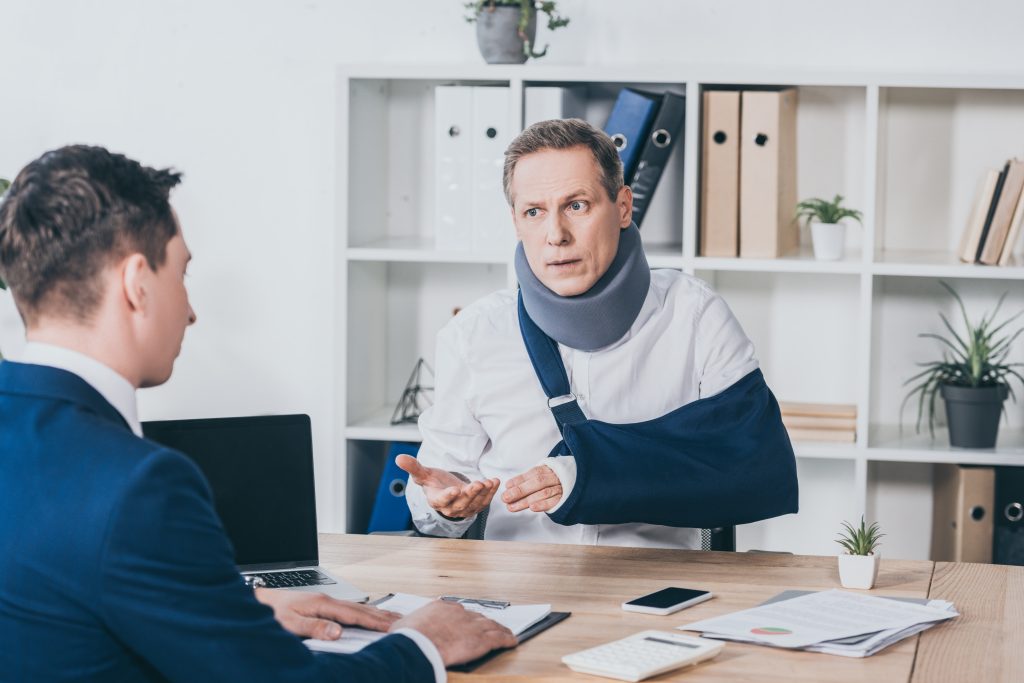 middle aged worker in neck brace with broken arm sitting at table and talking to businessman in blue