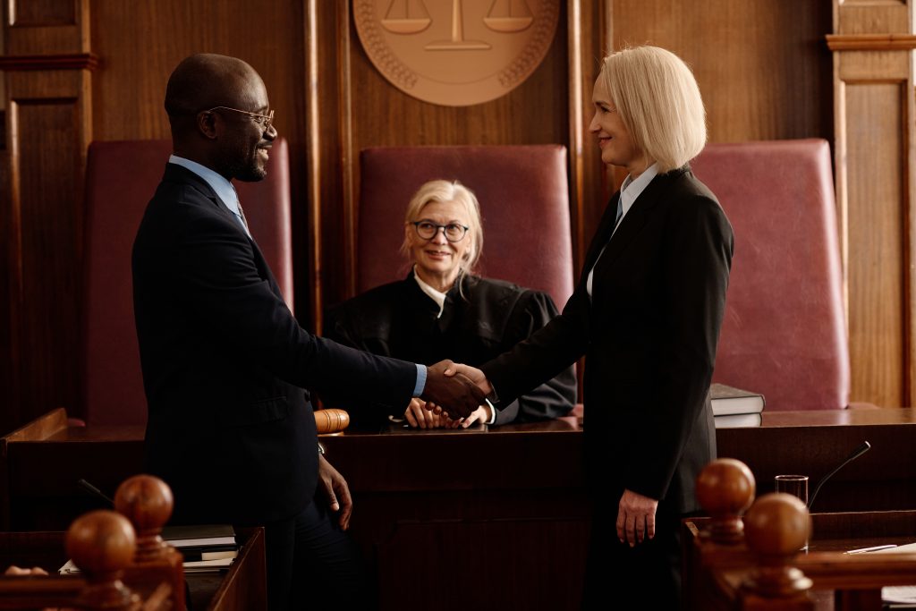 Side view of happy prosecutor and attorney shaking hands after making agreement against smiling mature female judge during trial session