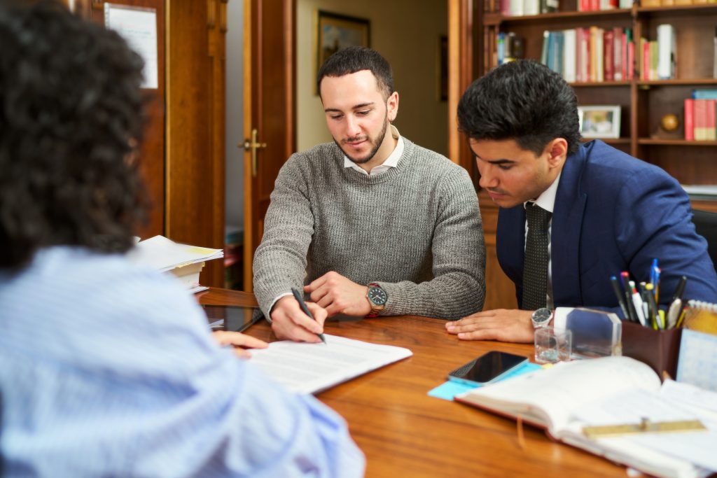 Three people are sitting at a table with papers and a pen. One of them is writing something down. Scene is serious and focused