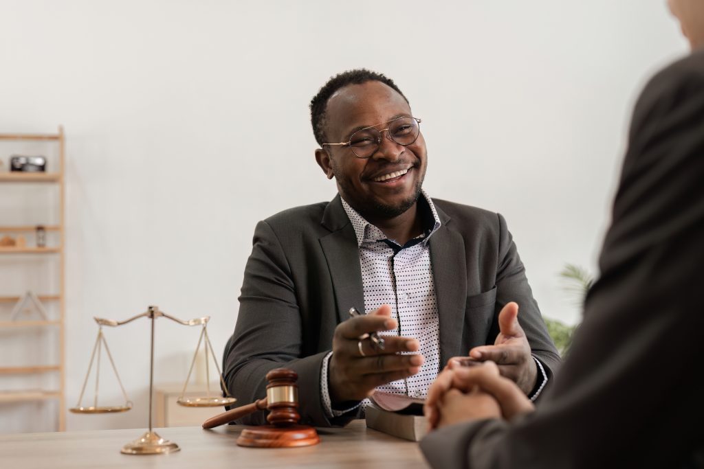 A man in a suit and glasses, likely the Best Attorney, is smiling and gesturing while seated at a desk, conversing with another person whose back is to the camera. A gavel and scales of justice are on the desk, suggesting a legal or professional setting.