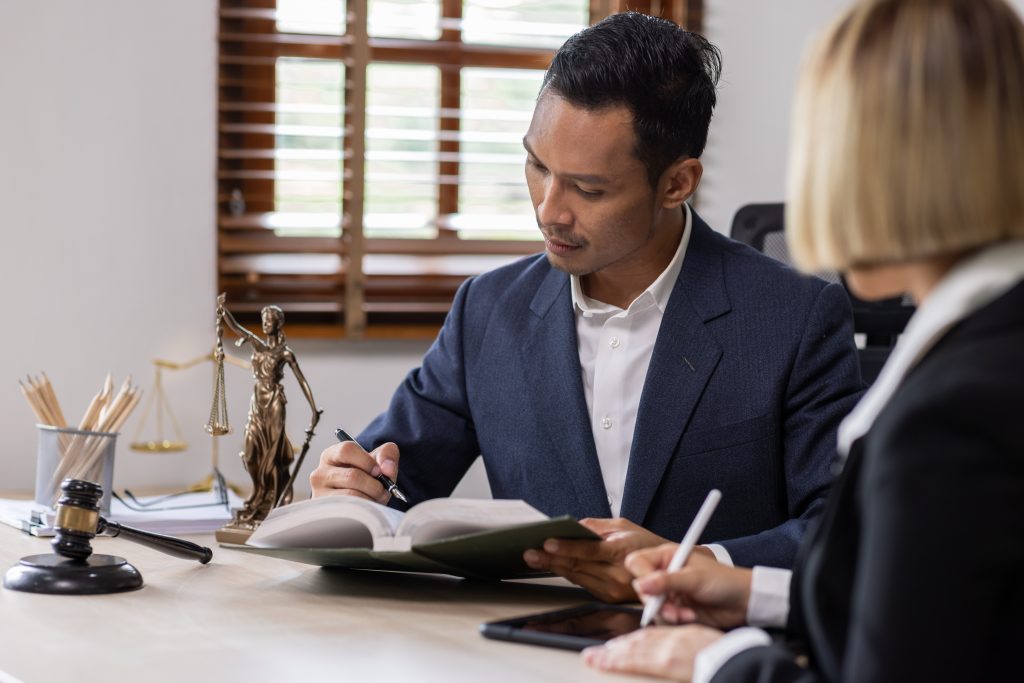 A man in a suit is writing in a notebook at a desk, with Lady Justice and a gavel nearby, likely discussing key differences in insurance policies. A woman sits beside him, also writing. The setting appears to be an office with a window in the background.