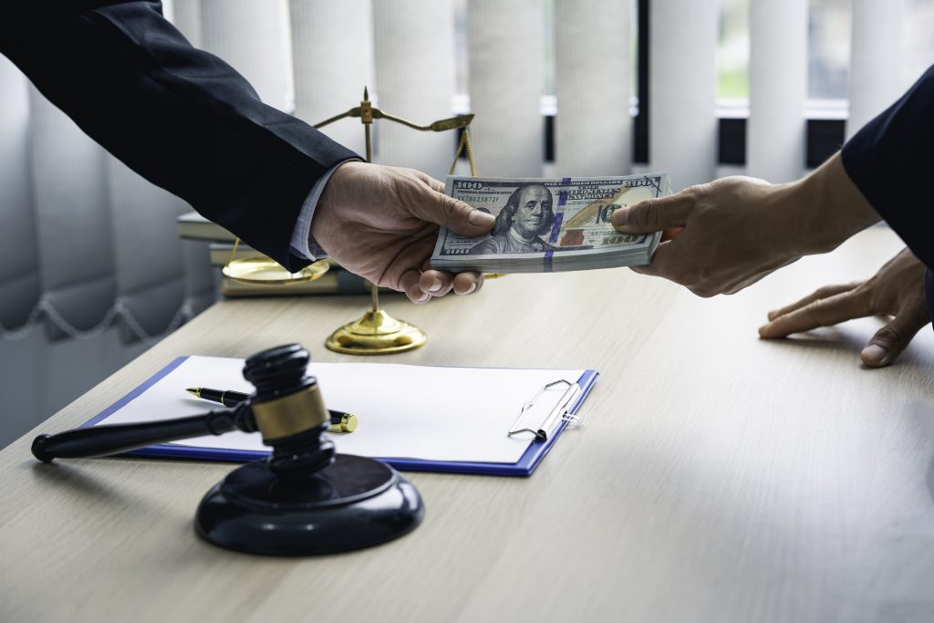 Two people in business attire exchange dollar bills across a desk. A gavel, scales of justice, a clipboard with a pen, and books are visible. The scene suggests an understanding of damage compensation in a legal or business transaction.