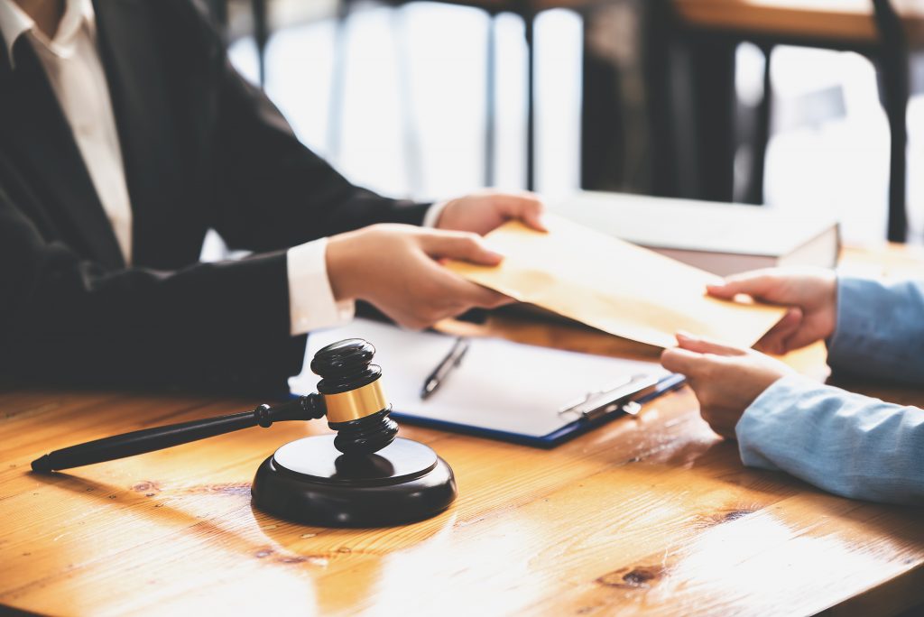 Two people exchange an envelope over a wooden table, with a gavel in the foreground. Papers and a book on the table suggest a legal or formal setting, possibly discussing filing tips for an insurance claim.