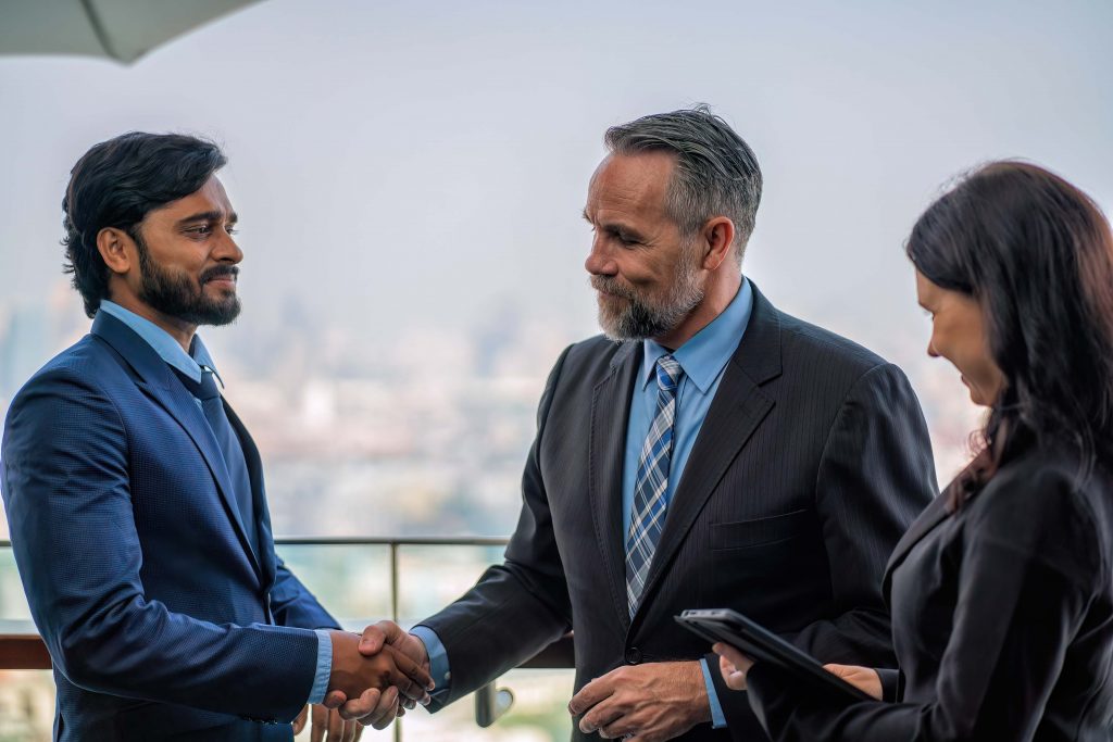 Three business professionals in suits are outdoors on a balcony with a cityscape in the background. As two men, possibly discussing first-party insurance, shake hands firmly, a woman with a tablet stands nearby, smiling and observing the handshake.