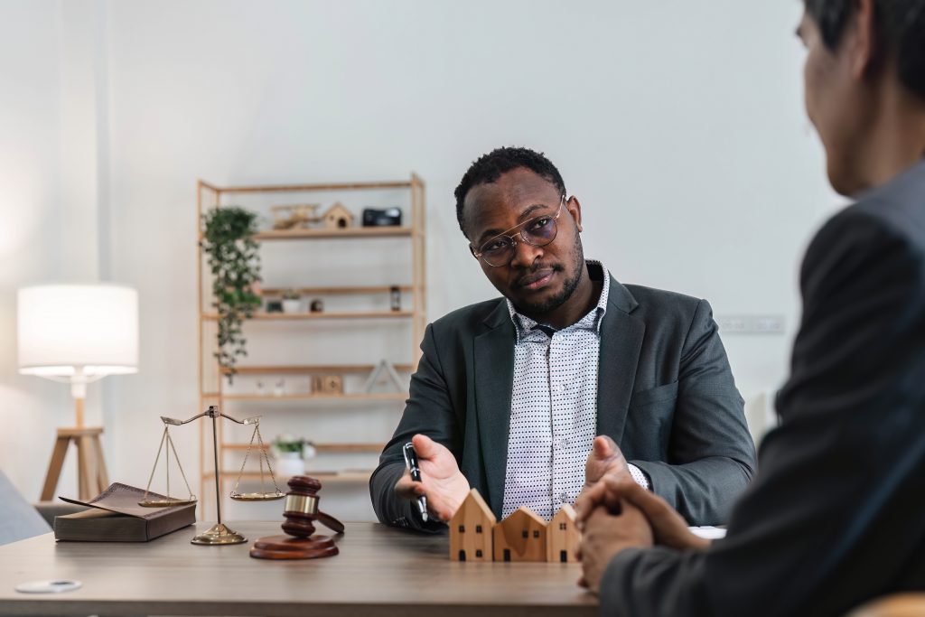 A man in a suit sits at a desk with scale models of houses, a gavel, and scales of justice. He seems to be discussing a property damage lawsuit with another person. Shelves and a lamp are in the background, suggesting an office environment.