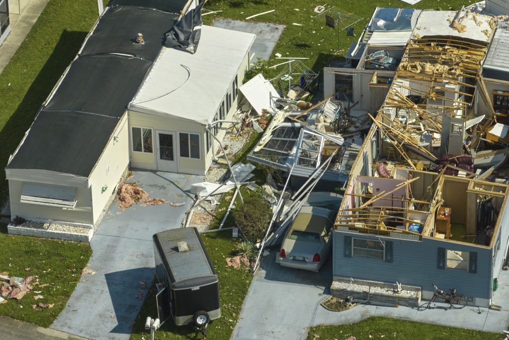 Aerial view of storm-hit properties reveals one house with a partially intact roof, while the other is heavily damaged, debris strewn about. A car rests in the driveway next to a small trailer. This significant property damage could potentially lead to a lawsuit for repairs and restitution.