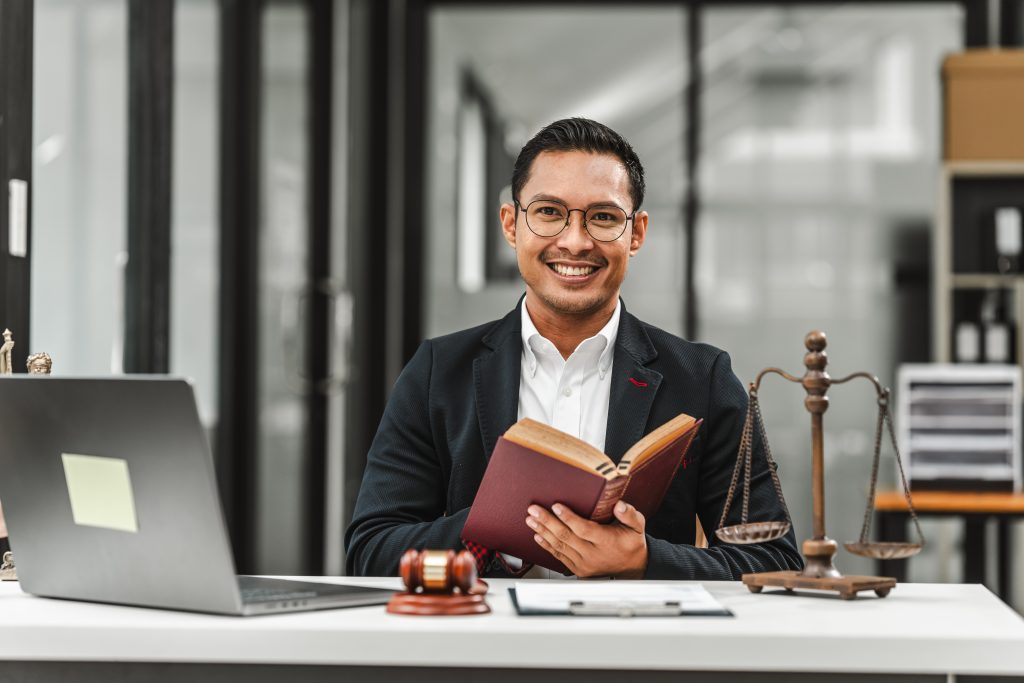 A man in a suit sits at a desk, smiling over an open book about the difference between suing and pressing charges. On the desk are a laptop, a gavel, and scales of justice. The background features bookshelves and large windows.