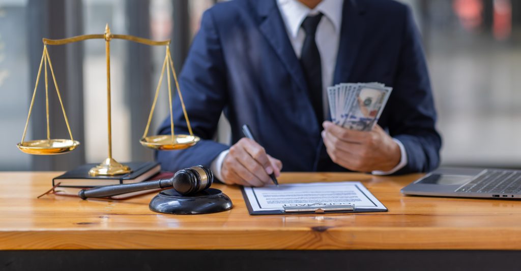 A person in a suit, holding a stack of dollar bills, sits at a desk with an essential guide and pen. Nearby are a gavel, a brass scale, and a laptop. The setting suggests an environment focused on damage compensation in legal or financial matters.