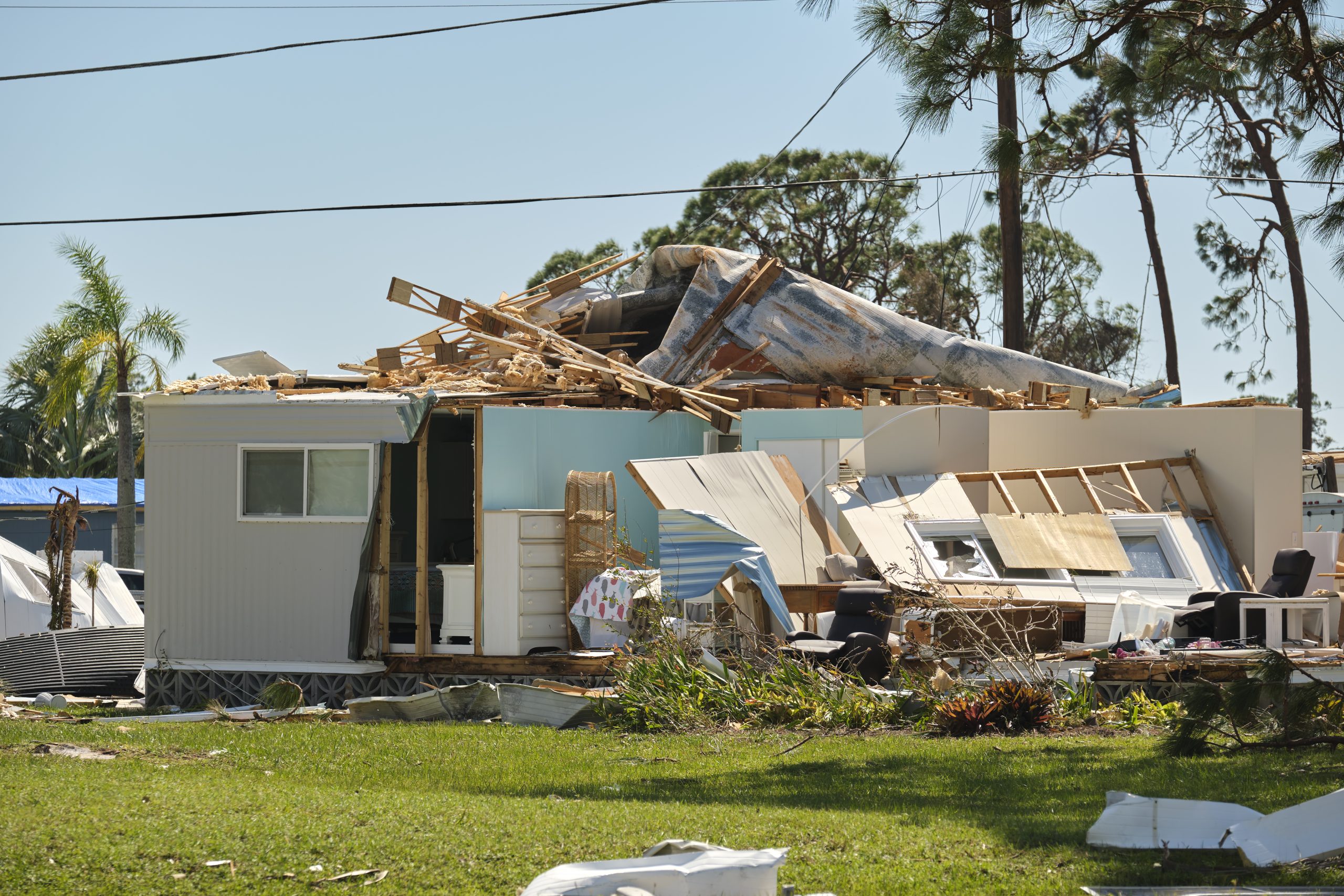 A damaged house with a collapsed roof and walls speaks volumes of property damage. Debris surrounds the building, and furniture is visible inside. Overhead power lines and trees are in the background under a clear sky, setting the scene for a potential lawsuit file.