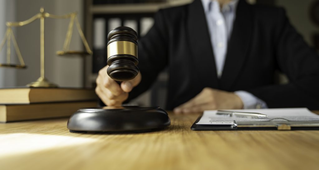 A person in a suit is seated at a desk, preparing to strike with a gavel. On the desk are a clipboard with papers, a pen, and law books. Scales of justice are visible in the background, highlighting the difference between pressing charges and suing in this legal setting.