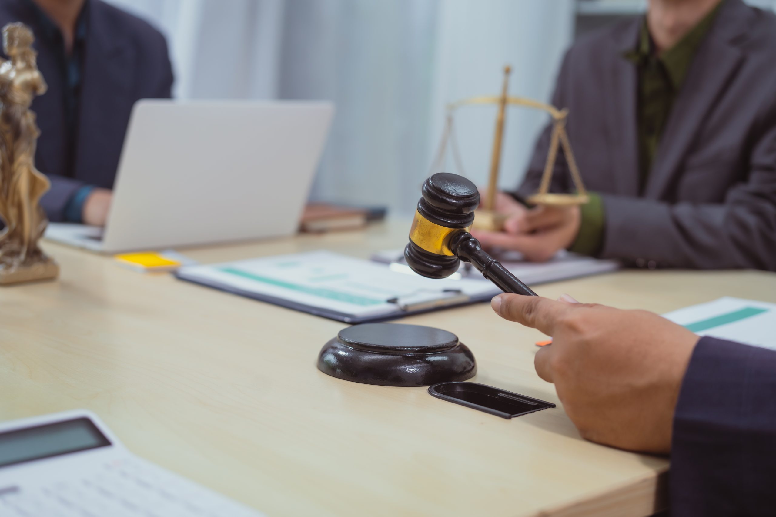 During the meeting, a person holds a gavel over the table, emphasizing the difference between pressing charges and pursuing a lawsuit. In the background, one individual is equipped with a laptop while another handles scales of justice. Documents and a calculator are scattered across the tabletop.