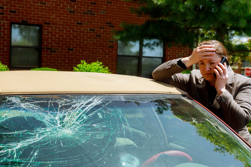 A woman stands by a car with a cracked windshield, holding a phone to her ear. She looks concerned, with one hand on her head. A brick building and trees are in the background as she considers filing tips for an insurance claim.