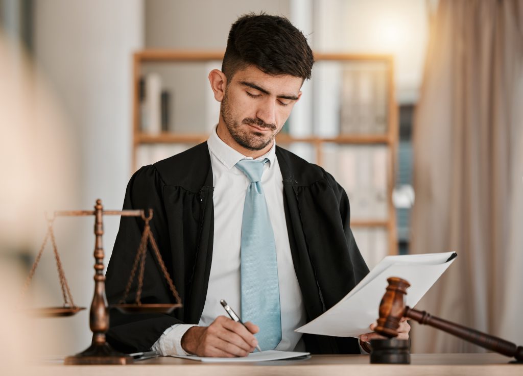 A man in a suit and tie, wearing a black robe, sits at a desk with a gavel and scales of justice. He is examining expunged documents and taking notes. The background features shelves with books.