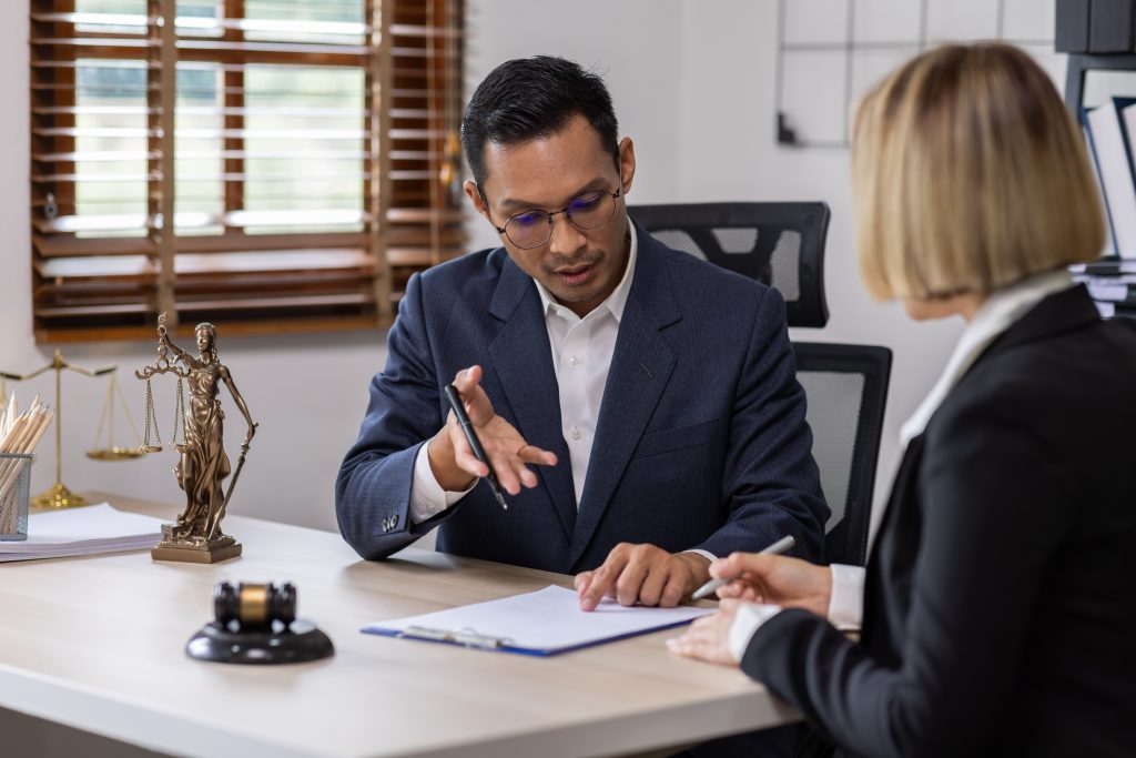 A man in a suit, holding a pen, discusses personal injury documents with an attorney in an office setting. The desk has a justice statue, a small gavel, papers, and an organizer. Window blinds are partially open, letting in natural light as questions arise.