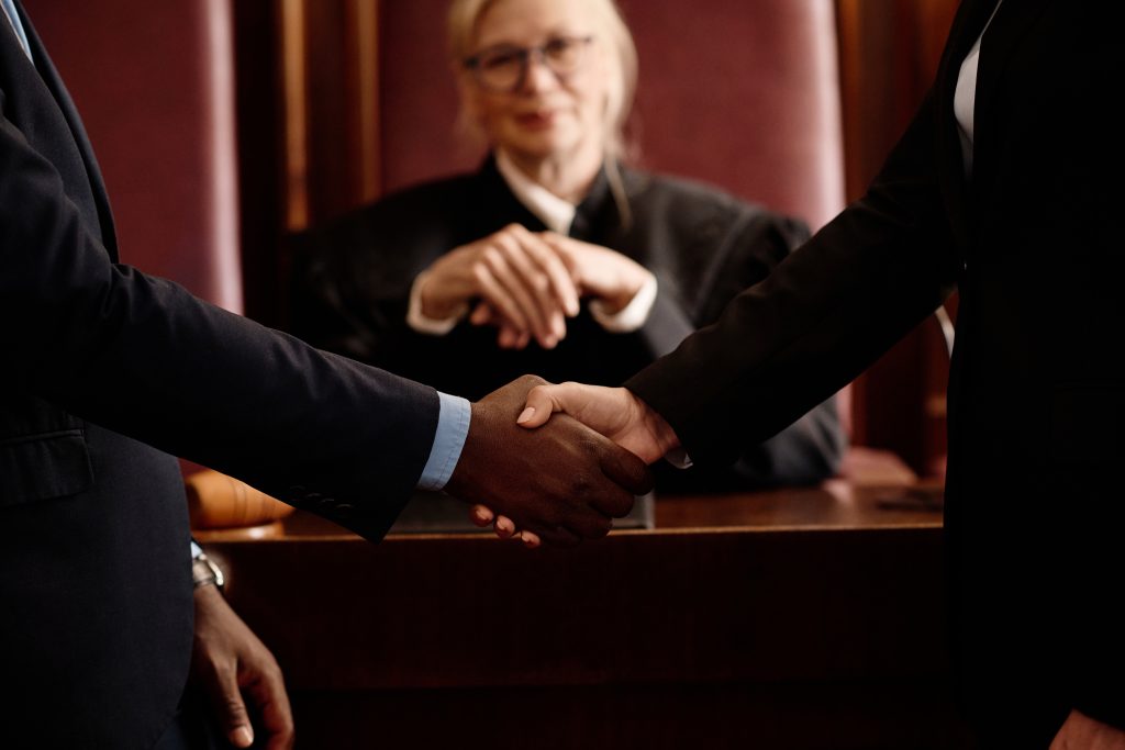 Two people are shaking hands in front of an attorney seated at a wooden desk in a courtroom. The judge, clad in a black robe, observes the exchange with the gavel nearby, indicating resolved questions in this personal injury case.