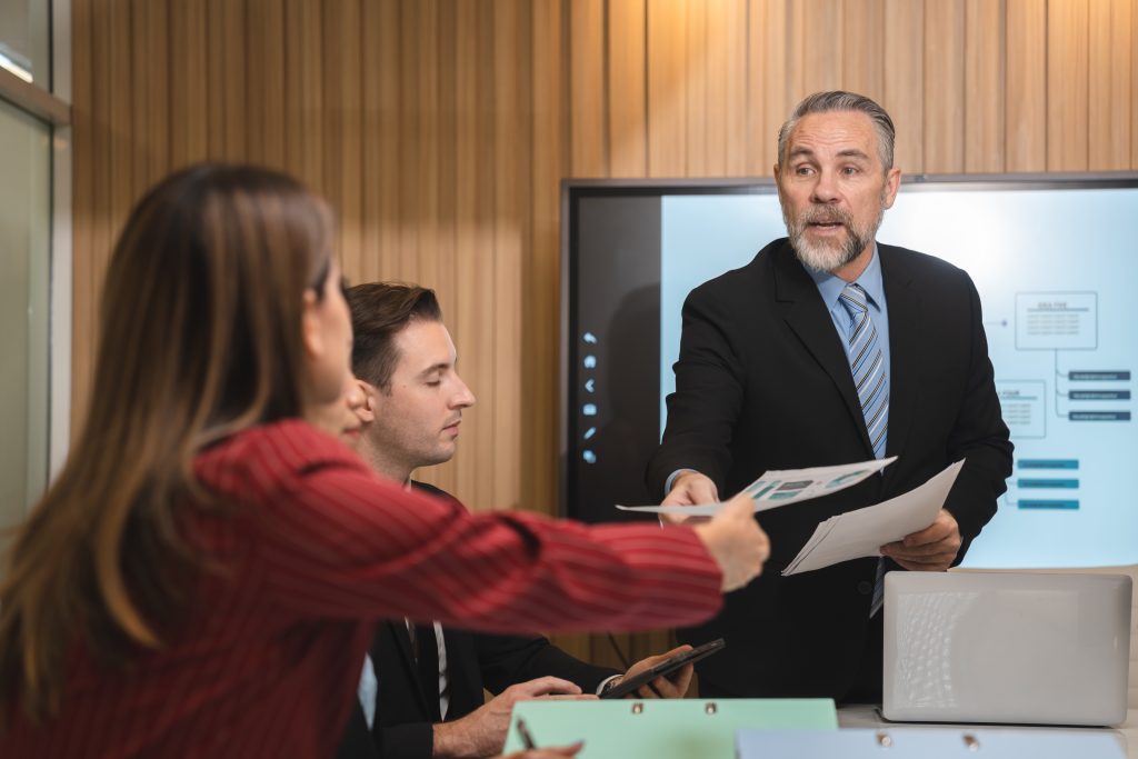 During a meeting, a man in a suit hands documents to a woman in a red blazer. Beside her, another man holds a tablet, while charts are displayed on the presentation screen. They discuss tips to win personal injury cases effectively and efficiently.