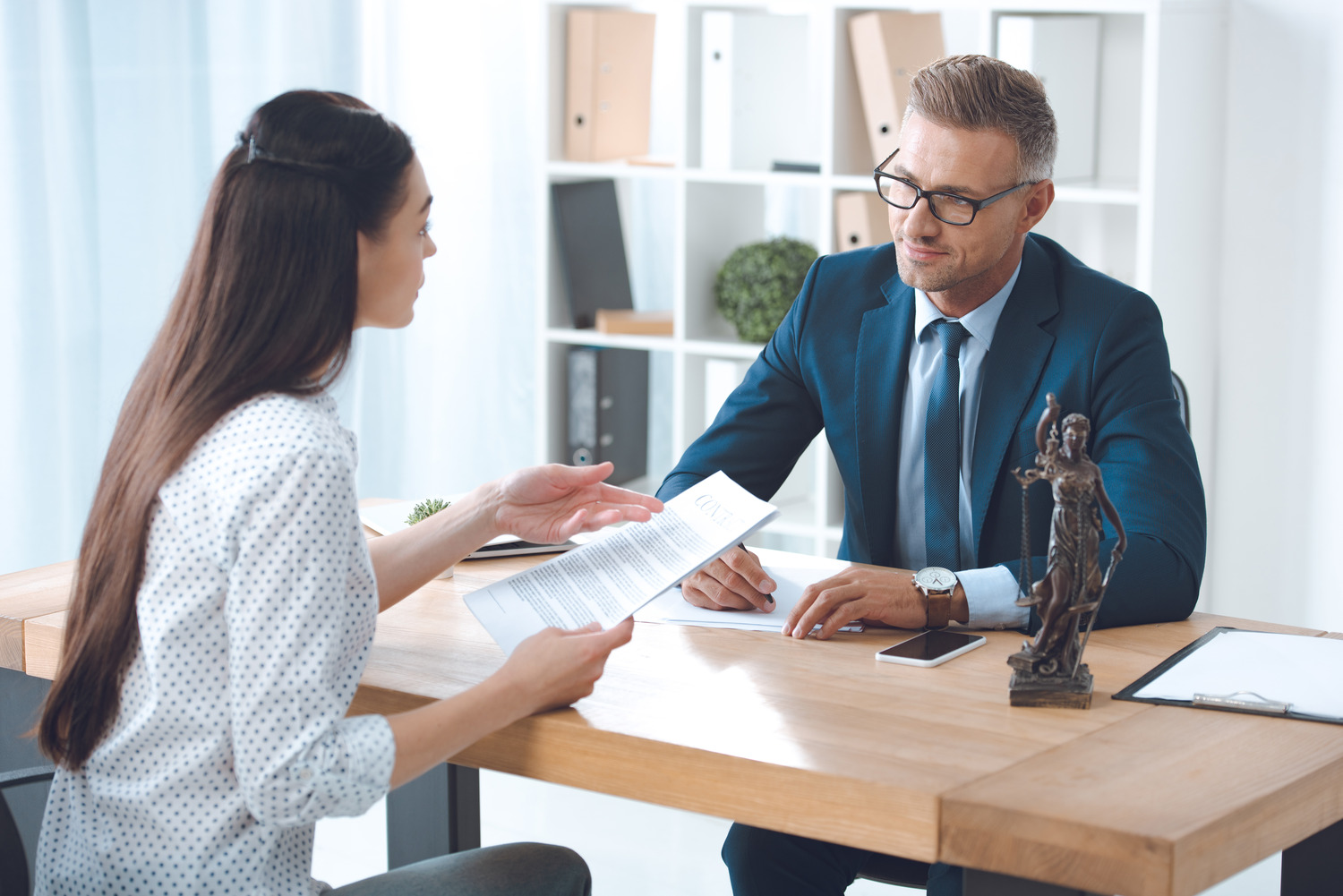 A woman and a man in formal attire sit across from each other at a wooden desk in a well-lit office. The woman, an attorney, holds a personal injury document, while the man listens attentively. A small statue is on the desk beside a clipboard and a phone.