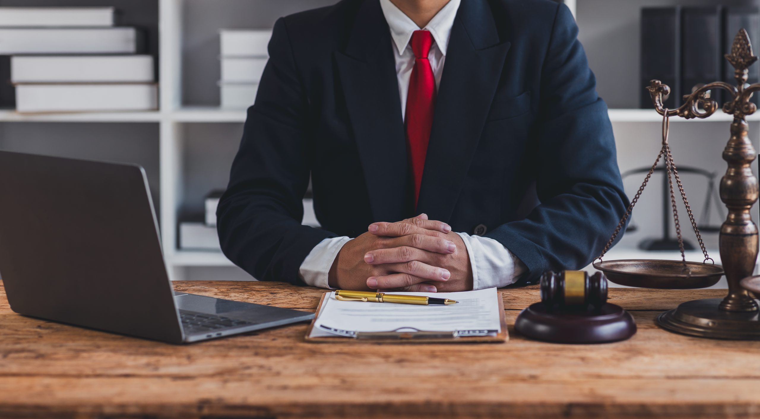 A person in a suit with a red tie sits at a wooden desk, contemplating the key differences in legal cases. In front of them are a laptop, a clipboard with papers and a pen, a gavel, and a set of scales. Shelves with boxes fill the background.